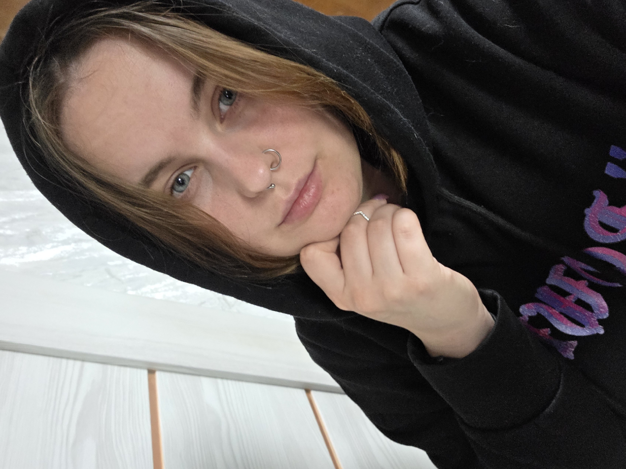 A close-up, tilted, eye-level shot depicts a person with fair skin and light brown hair partially obscured by a black hooded sweatshirt. The individual has blue eyes and multiple piercings on the nose. The left hand supports the chin; the hand has a ring. The background includes a white wall or surface, along with thin vertical support. The location is unknown.