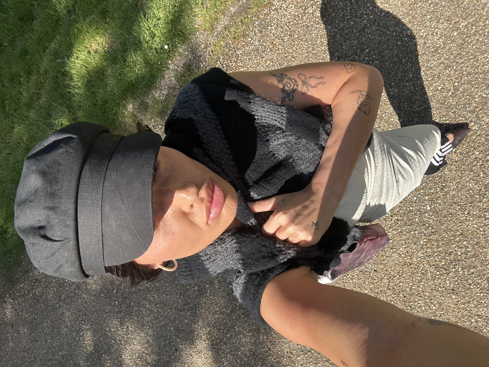 A top-down, eye-level shot depicts a person posing outdoors on a paved surface, likely a path or driveway. Patches of grass are visible in the upper left corner. The individual is wearing a dark, wide-brimmed hat, a grey and black knit top, and light-colored trousers. One shoe, with black and white stripes, is partially visible. The person's arms are visible, adorned with tattoos. They are touching their top near the chest. The shadow of a person and camera can be seen on the ground, suggesting the photo was taken by the person themselves. The setting is likely a residential area in Tunbridge Wells, United Kingdom, based on the typical British grass and stone surface.
