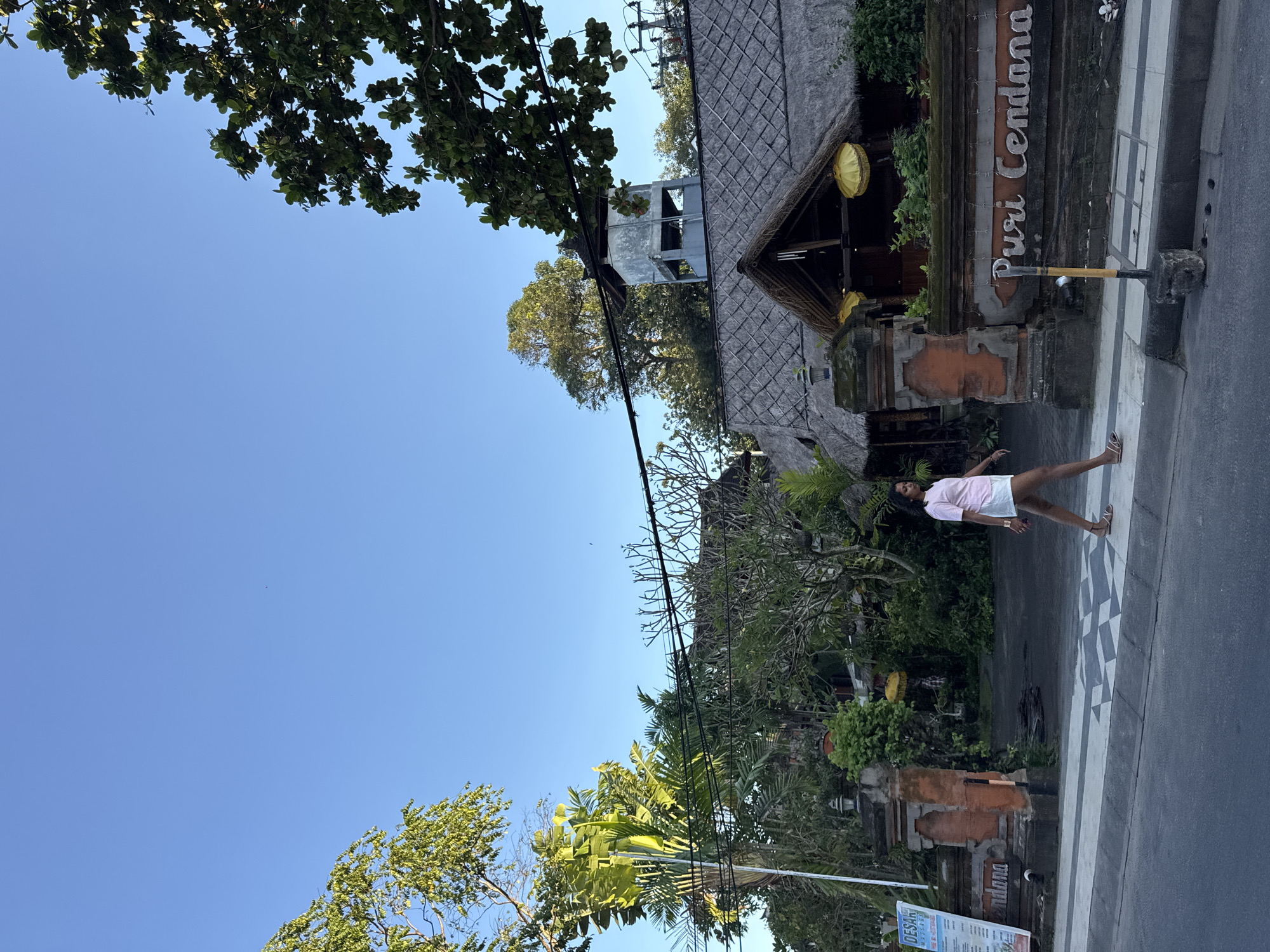 A daytime image depicts a section of road in Kuta, Indonesia, with a person standing on a decorative crosswalk in front of a building. The person is light-skinned, wearing a light pink top and light-colored shorts. The building is marked with "Puri Cendana" on the facade. The architecture includes thatched roofing and Balinese-style stonework. Lush greenery, including trees and palms, surrounds the building. Overhead power lines stretch across the frame. The sky is clear and blue.