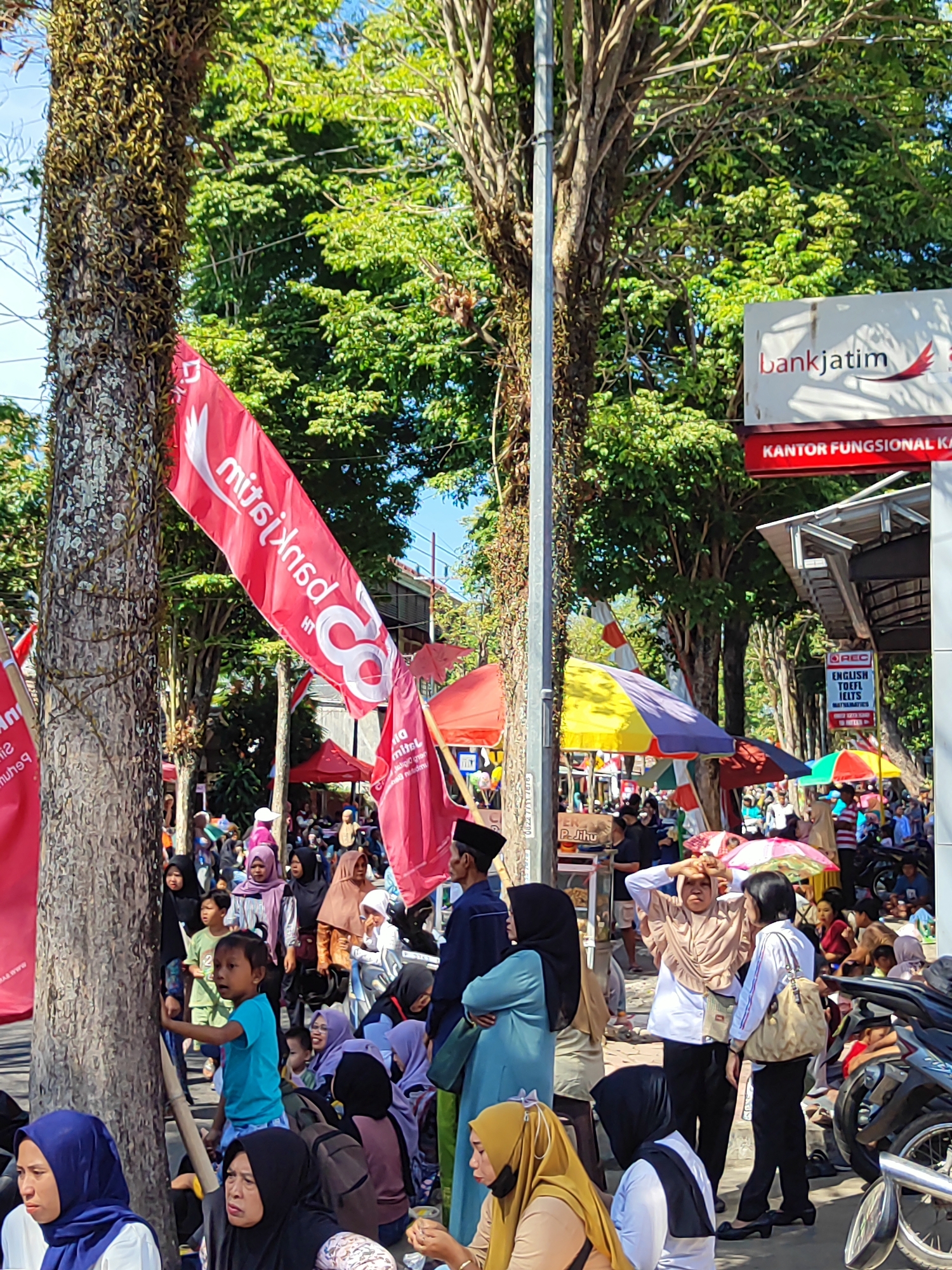 The image depicts a street scene in Dabasah, Indonesia, likely during an outdoor event or gathering. A prominent red flag with white text, partially obscured by a tree, is visible. In the background, a building with a "bankjatim" sign and "KANTOR FUNGSONAL KA" text indicates a bank branch. Several people are present, with many wearing headscarves, suggesting a predominantly Muslim population. Some individuals are standing, while others are seated or in conversation. Colorful umbrellas are scattered throughout the scene, providing shade. A motorcycle is parked on the right side of the image. The environment is characterized by trees and a clear sky, suggesting it is daytime. The overall impression is of a busy public space.