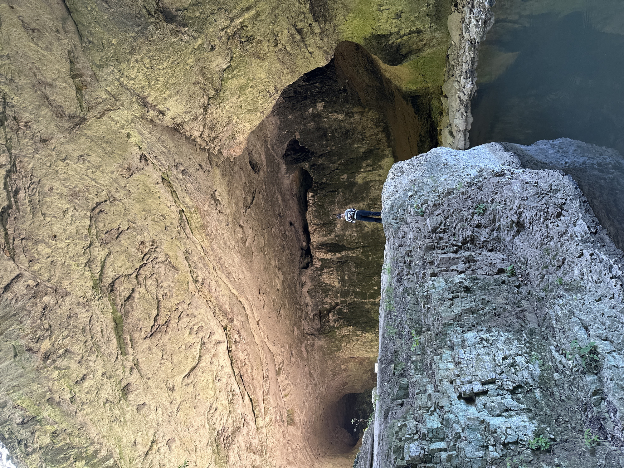 The image shows the Devetashka Cave, located near the city of Lovech, Bulgaria. The perspective is from inside the cave looking upwards, showing the cave's interior structure and large openings in the ceiling. A person is visible wearing climbing safety equipment, standing on a rock formation. The walls of the cave are textured, with visible layers of rock and signs of moisture.
