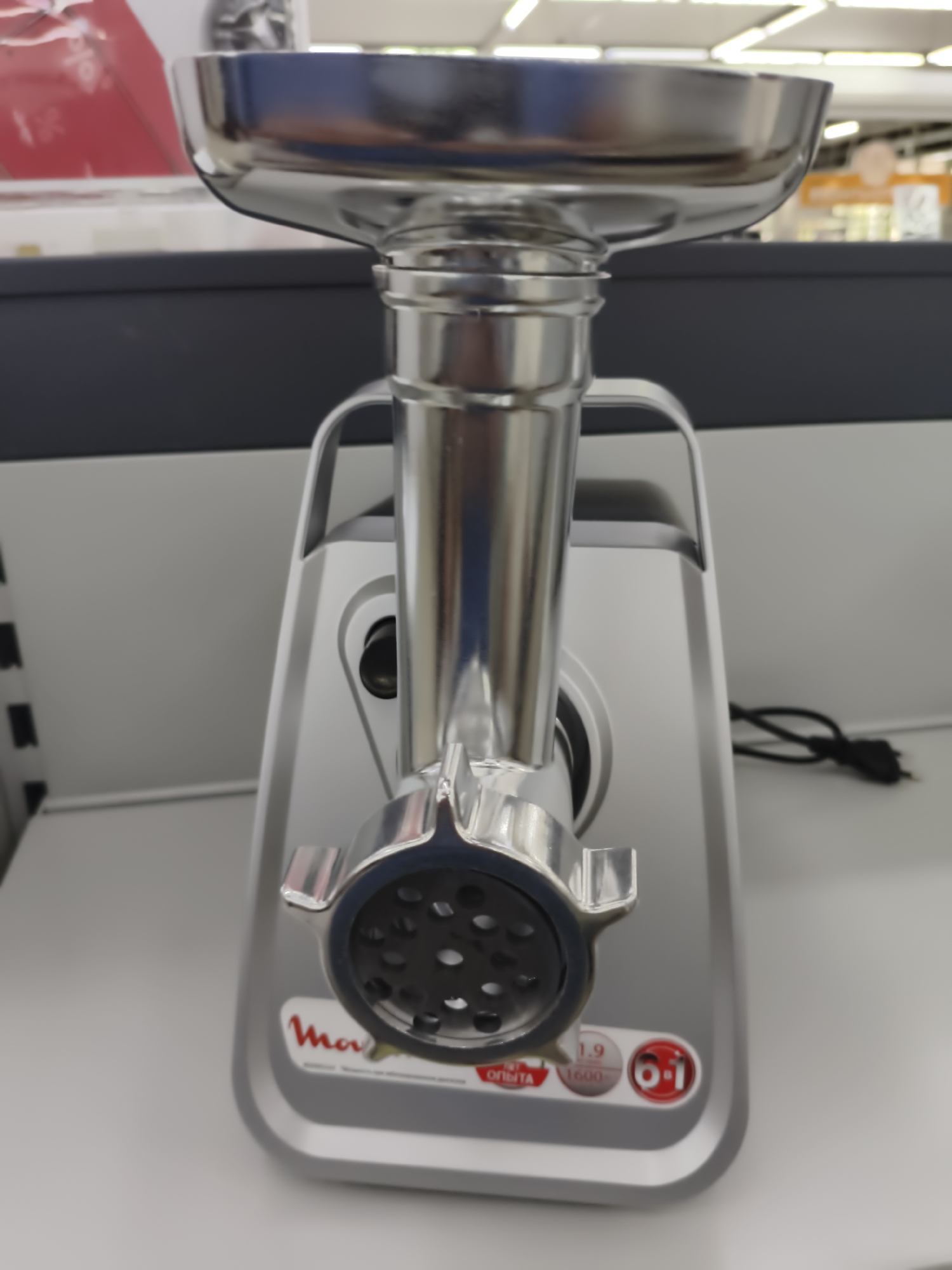 A silver meat grinder is positioned on a white shelf in what appears to be a retail environment. The meat grinder features a metal funnel at the top, a cylindrical metal grinding mechanism, and a base with various labels. One prominent label displays the brand "Moulinex" with accompanying text and circular icons. The background consists of shelving and what looks like store lighting. The scene is indoors and well-lit. There is a black power cord visible on the right side of the image. The overall impression is that of a product displayed for sale. The image does not provide any information about the city of Aktobe, Kazakhstan.