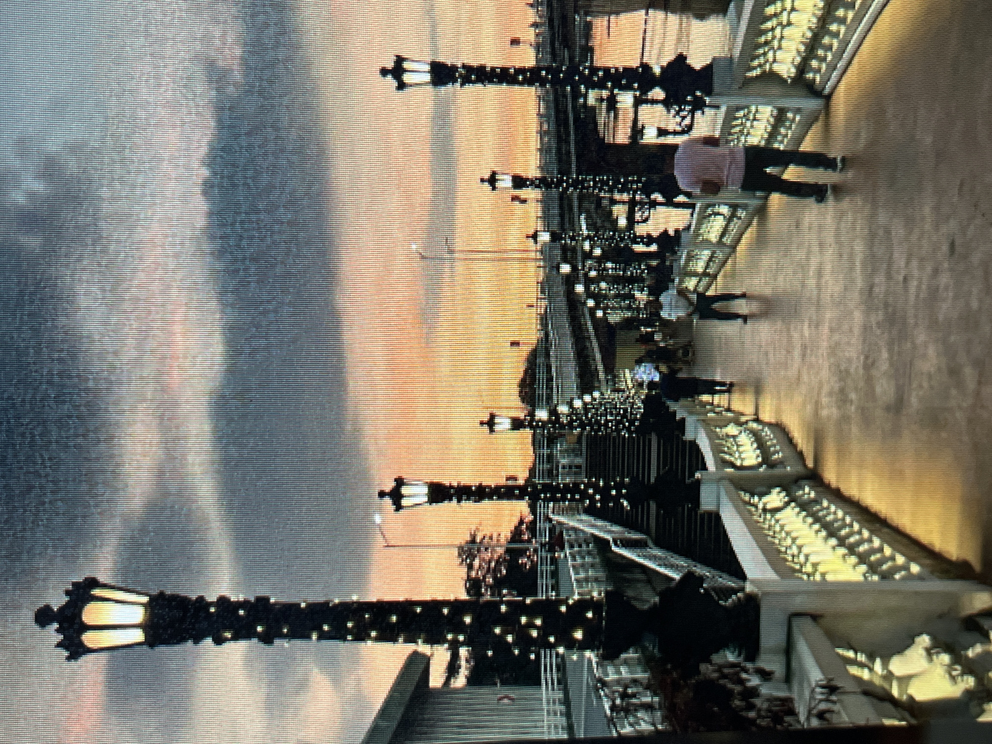 The image depicts a walkway illuminated by lampposts adorned with decorative lights. The sky exhibits a sunset with hues of orange and pink, contrasted by dark clouds. Several people are visible walking along the path. In the foreground, a white ornate railing lines the walkway, and to the left, a staircase leads up to the walkway. In the background, a bridge spans across a body of water. The scene appears to be a public space in the city of Sejahtera, Indonesia, possibly a waterfront promenade or park.