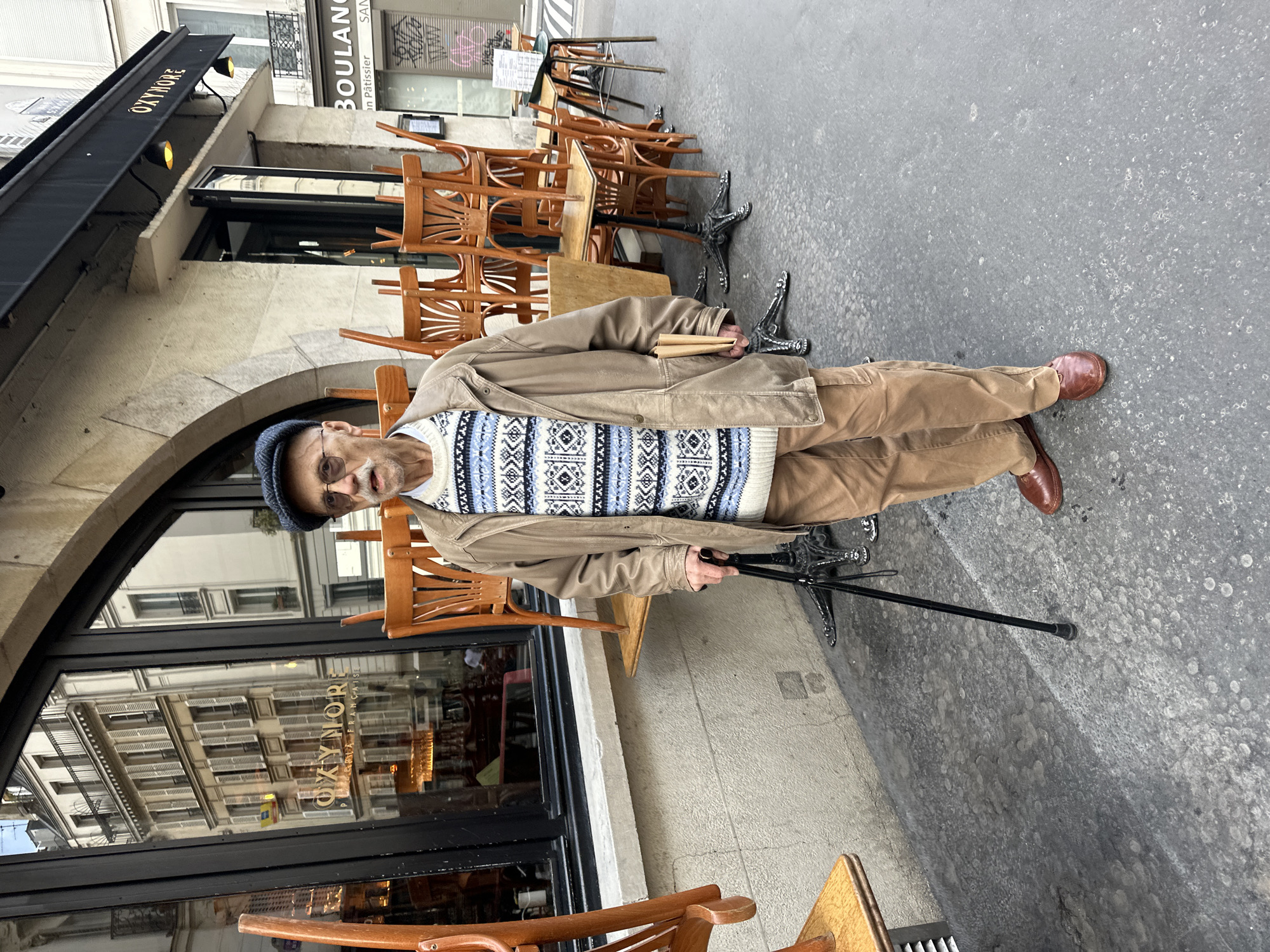 A man stands in front of a Parisian cafe. The cafe is named "Oxymore, Pâtisserie Française". In the background is a boulangerie. The man is holding a walking stick. Tables and chairs are stacked and standing outside the cafe. The street is paved.