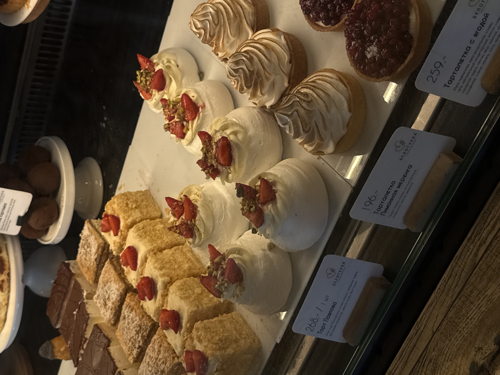 The image shows a display case filled with various pastries in a store, possibly a bakery or café. The pastries include:
* Tarts with berries on top
* Meringue-based desserts topped with strawberries and pistachios.
* Individual cake slices.
* A bowl with chocolate desserts.
Each set of pastries has a small label in Russian displaying the name of the dessert and price. The labels include brand name “Белотурка” (Beloturka). The image was likely taken in Russia, and the mention of "Beloturka" and the types of pastries sold here suggest this could be a cafe in the city of Izhevsk, Russia.