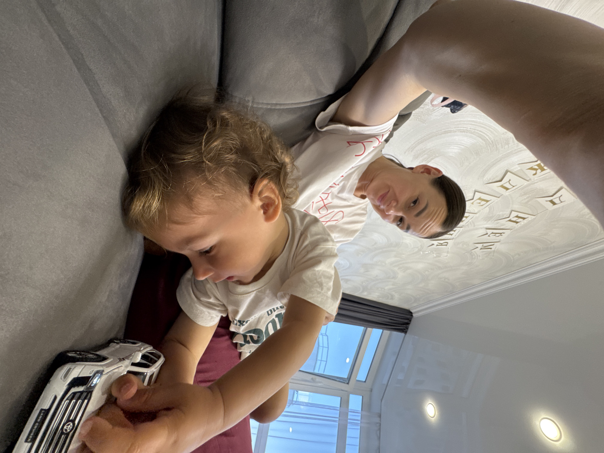 The image depicts an indoor scene in what appears to be a residential living space in Sochi, Russia. An adult woman with dark hair pulled back is positioned in the upper right, looking directly at the camera with a slight smile. She wears a white t-shirt with red Cyrillic text. Her right arm extends into the upper left, suggesting she is holding the camera to take a selfie.
Below and in front of the woman, a young child with light, curly hair is lying on a grey upholstered couch. The child, also wearing a white t-shirt, is oriented downwards and to the right, focused on a white toy model car, identified as a "Land Cruiser," held in their hand. A dark red cushion or upholstery is visible beneath the child.
The background features a wall with a subtle patterned wallpaper, adorned with white banner-style decorations displaying golden Cyrillic letters that spell out portions of "С ДНЕМ РОЖДЕНИЯ" (Happy Birthday). The ceiling is a white, reflective surface, fitted with two active circular recessed lights emitting a warm glow. A large, multi-paned window is visible on the right, showcasing a clear blue sky and an indistinct outdoor structure, with dark grey curtains drawn partially across it. The scene is brightly lit, indicative of daytime.