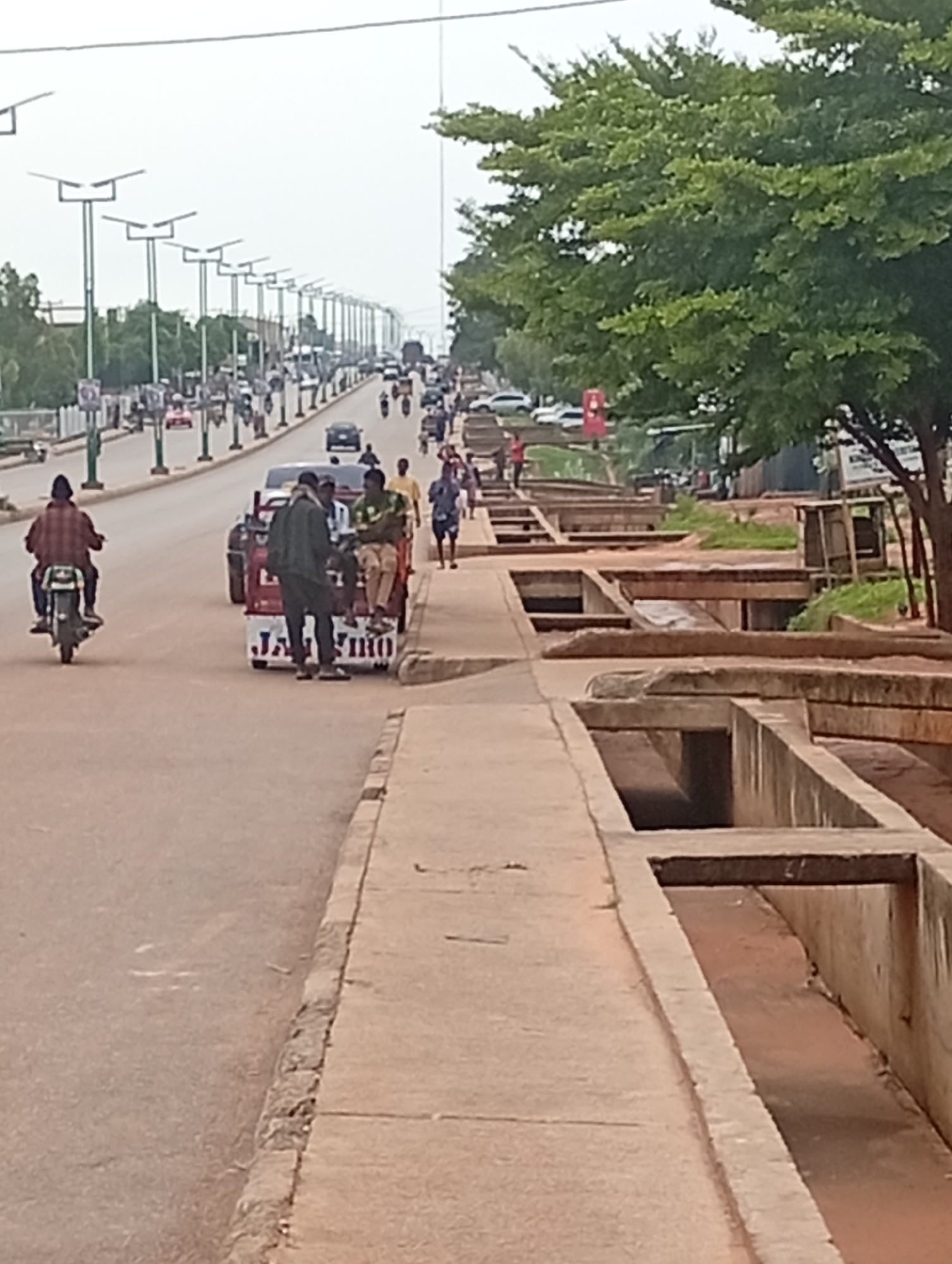 The image depicts a street scene in Tumfure, Nigeria. In the foreground, a concrete sidewalk runs parallel to a paved road. A channel or drain runs alongside the sidewalk. Several people are visible on the road and sidewalk. A person is riding a motorcycle on the left side of the road, facing away from the camera. In the center, a three-wheeled vehicle, possibly a type of motorized rickshaw, is stopped on the road. Several people are standing around or sitting in this vehicle. Further down the road, there is traffic, including cars and motorcycles. The street is lined with streetlights on both sides. On the right side of the image, a large tree with green foliage is prominent. The sky is overcast. The overall impression is a busy urban street with pedestrian and vehicular activity.