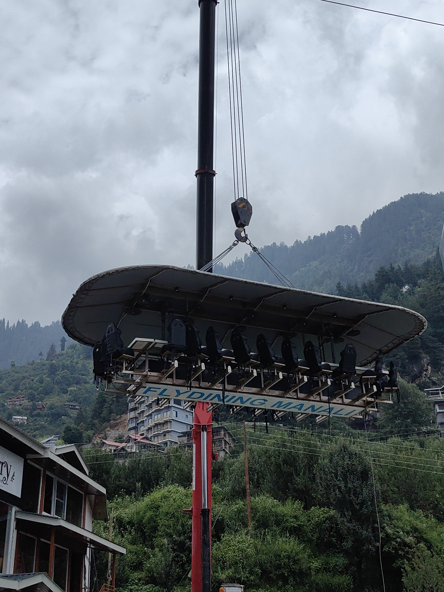 The image depicts a unique dining experience known as "Dinner in the Sky" or a similar elevated restaurant setup. A large, circular platform holding multiple seats around a central table is suspended in the air by a crane. The platform is dark gray and appears to be made of metal and fabric. Black seats with safety harnesses are arranged around the perimeter of the platform.
Below the platform, a red crane arm is visible, supporting the structure. Text on the underside of the platform reads "FLYING MANALI".
The background shows a lush, green mountainside with dense trees. Several buildings, including a multi-story structure with balconies, are visible on the slopes. The sky is overcast with clouds.
The location is identified as Vashisht, India, a town near Manali known for its hot springs and scenic mountain views. The scene suggests an adventurous and unconventional dining opportunity, capitalizing on the picturesque landscape. There are no people clearly visible on the platform, indicating it may be set up or in the process of being prepared for operation.
