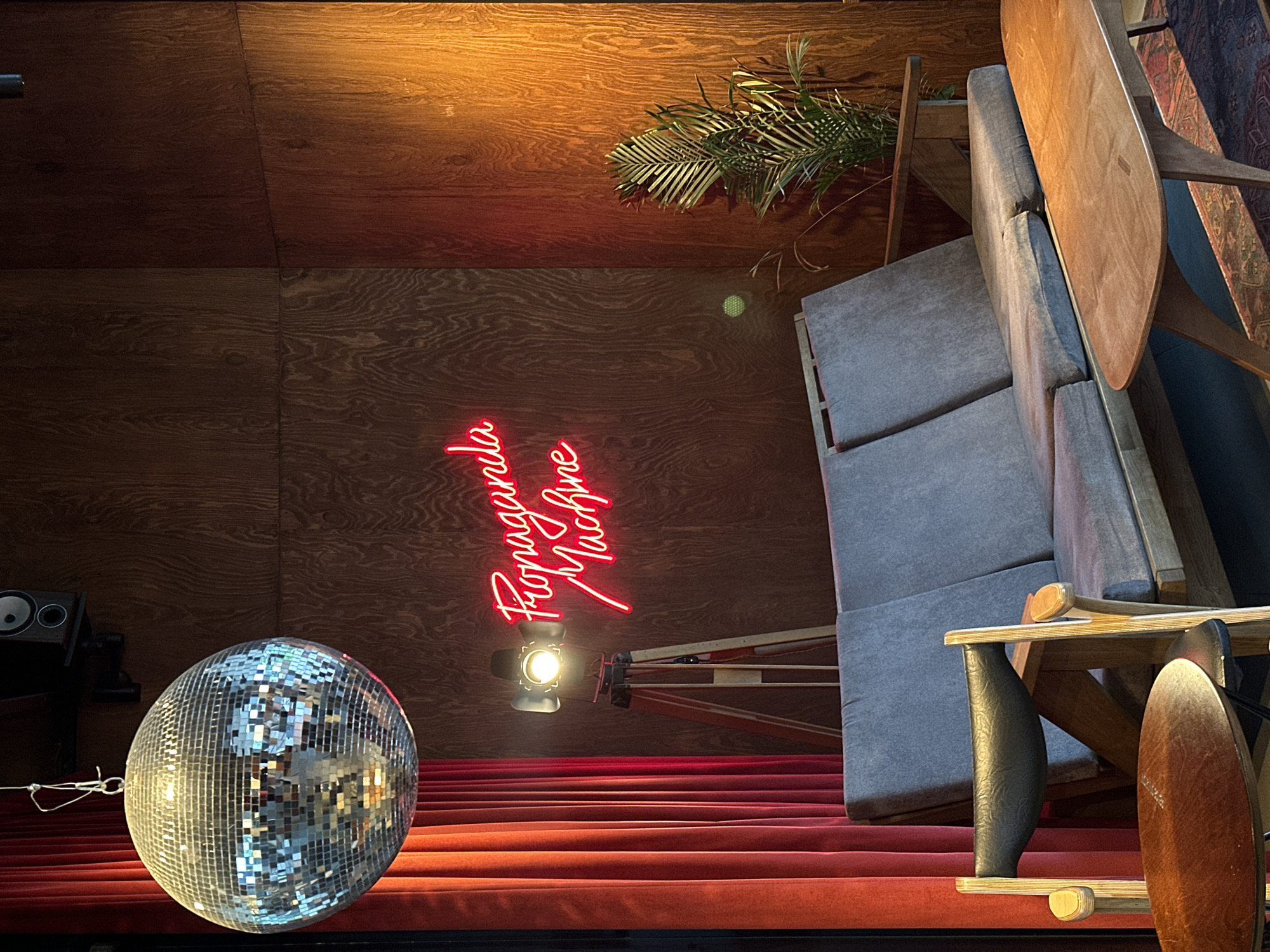 This image captures a unique scene at Propaganda Machine, a venue located in Unknown, Unknown. The backdrop features wood-paneled walls, partially illuminated by a red neon sign that spells out "Propaganda Machine." A disco ball hangs prominently in the foreground, reflecting light across the scene. A stage-style setup with red carpeting is visible, along with a spotlight directed towards the audience area. Furniture, including a gray sectional sofa and a wooden table, is positioned off to the side. A potted fern adds a touch of greenery to the space. The overall atmosphere is reminiscent of a lounge or entertainment venue.