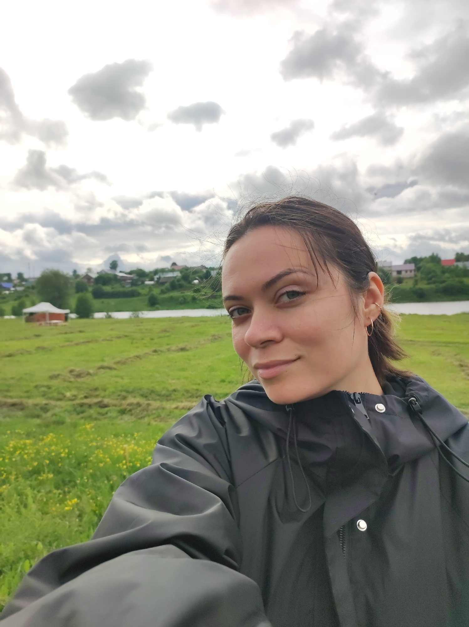 A young woman is the central subject of this outdoor selfie, taken in a rural or semi-rural setting identified as Фоки, Russia. She has dark hair pulled back from her face, light skin, and gazes directly at the camera with a calm, closed-lip smile. She is wearing a dark, possibly black or charcoal grey, waterproof jacket or windbreaker, featuring silver snap buttons and visible drawstrings around the neck. In her left ear, she sports a small gold hoop earring and an additional twisted wire earring higher on the cartilage.
The background of the image presents a lush, expansive green field, scattered with patches of small yellow wildflowers, likely dandelions. Beyond this field, a body of water, possibly a river, meanders through the landscape. On the opposite bank of the water, a village or cluster of houses with various colored roofs, including several prominent red ones, is visible amidst trees and foliage. To the left side of the frame, within the green field but closer to the water, a distinctive light-colored gazebo-like structure with red supports can be seen.
The sky is heavily overcast with a mix of white and grey clouds, suggesting a cloudy day, potentially cool weather given the woman's jacket. The scene conveys a tranquil, natural environment, characteristic of a countryside or riverside village. There is no visible text in the image.