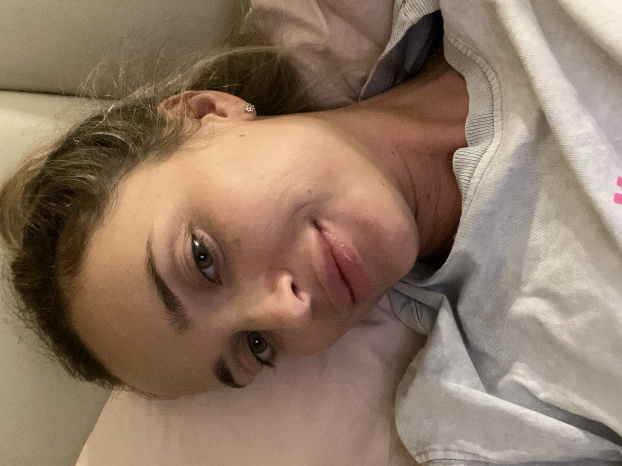 A close-up, overhead shot shows a woman lying on her side, facing the camera. She has light skin, brown hair pulled up in a ponytail, and green eyes. Her mouth is slightly open, and she is wearing a light blue t-shirt. A pink pillow is visible behind her head. The scene appears to be indoors, possibly a bedroom, and the lighting suggests it is nighttime. There is no indication of the location being Sharm El Sheikh, Egypt, or any other specific geographical area.
