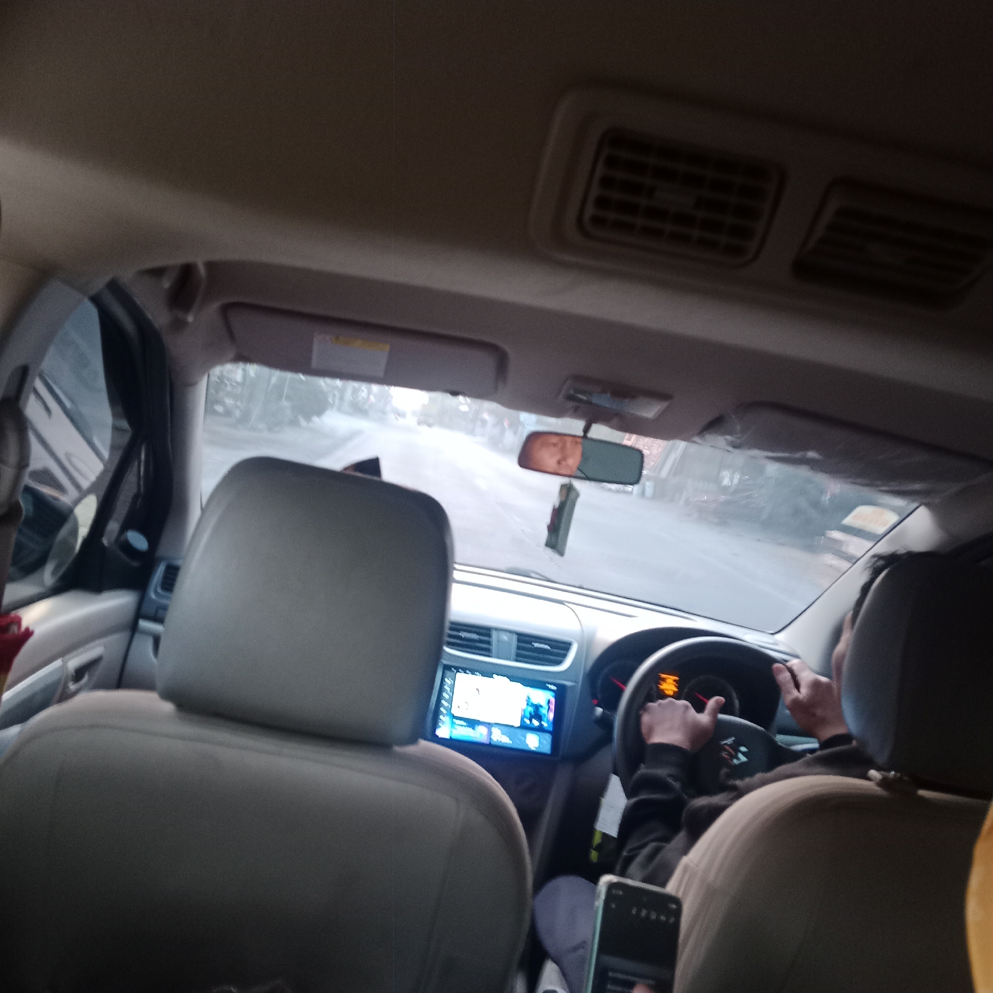 The image shows the interior of a car, likely taken from the back seat, looking forward. The driver, wearing a dark long-sleeved shirt, is visible from behind, holding the steering wheel. A car's dashboard with a display screen and air vents are prominent. The car appears to be in motion on a road, with a blurry view of trees and buildings visible through the windshield. A rearview mirror hangs from the ceiling, reflecting a portion of the driver's face. A mobile phone is visible in the foreground, held by someone in the back seat. The overall lighting suggests it might be daytime, possibly overcast or dusk. The location is identified as Tegowanu Wetan, Indonesia.