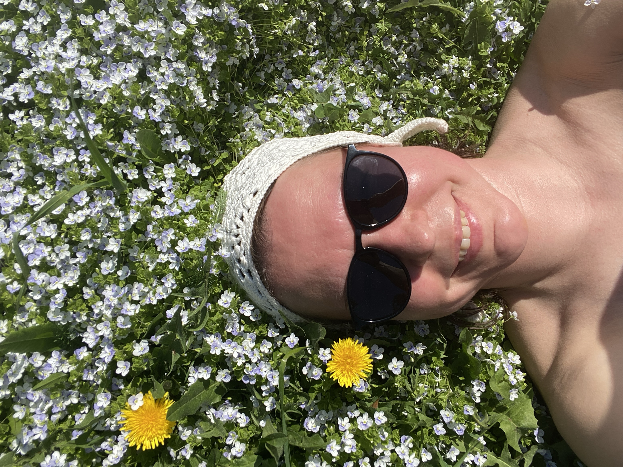 A woman is lying on her back in a field of small white and lavender flowers and a few dandelions. She is wearing a white crocheted cap and black sunglasses. The location is the city of Прикамский, Russia.