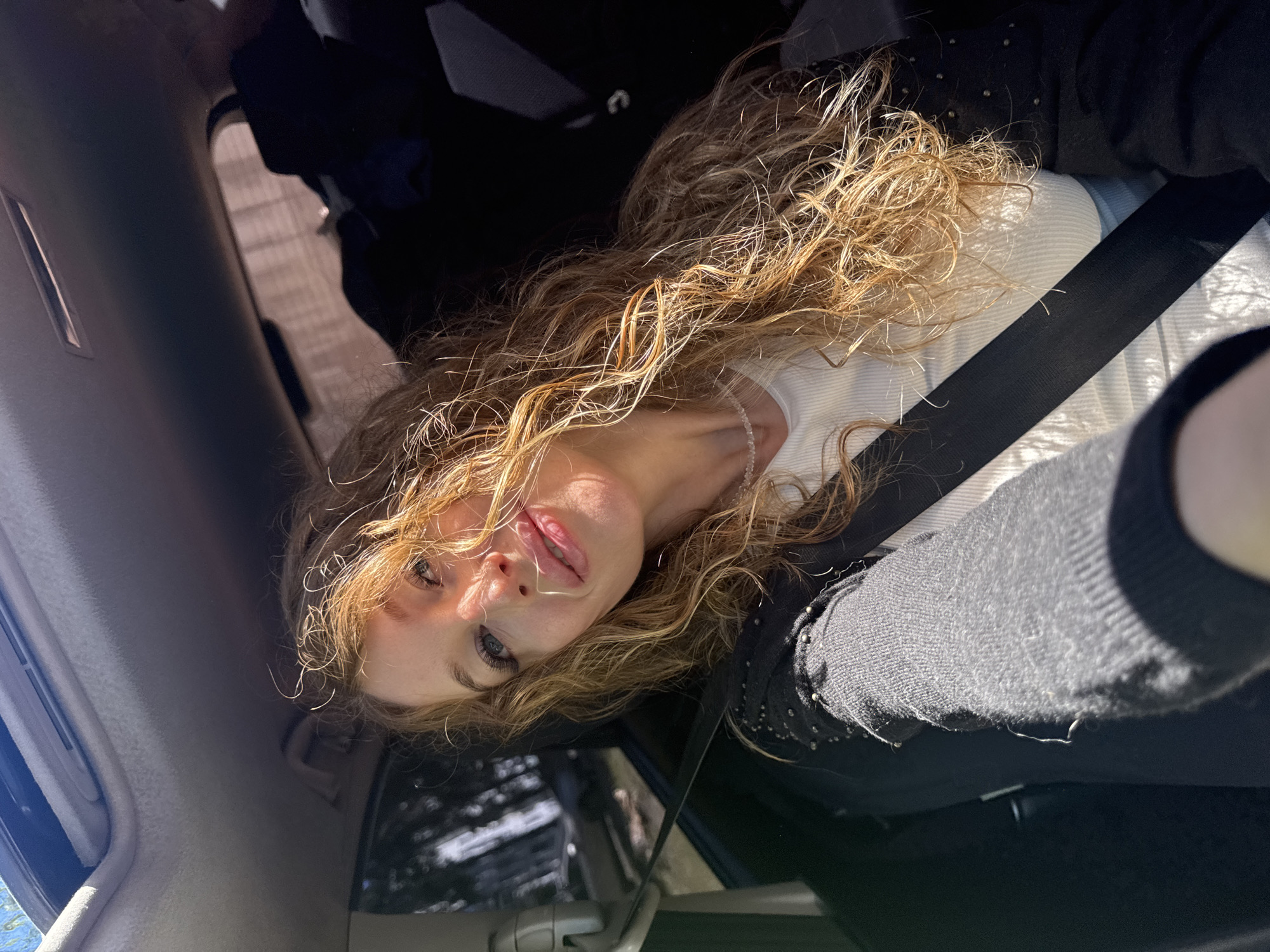 A woman with long, curly blonde hair is sitting in the passenger seat of a car, looking towards the camera. She is wearing a white ribbed top and a black cardigan adorned with small studs. A black seatbelt is visible across her chest. The car's sunroof is open, allowing sunlight to stream in, illuminating her face and hair. The background is blurred, showing the interior of the car and a glimpse of trees and a fence outside. The image appears to be a selfie taken on a sunny day. There is no information in the image to suggest the location is in the city of Chaikovsky, Russia.