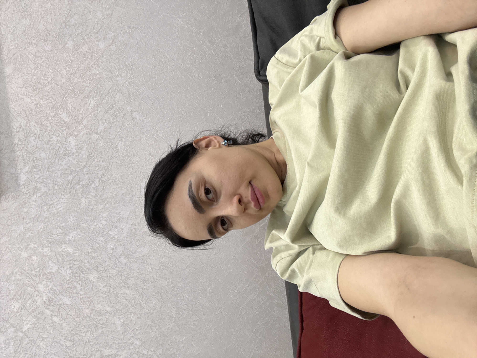 The scene is an interior setting within what is indicated as the city of Tbilisi, Georgia. A single adult female is depicted seated on a sofa. The sofa includes a dark red cushion on the left and a dark gray cushion positioned behind the individual. The background wall is light gray with a textured finish. The female has dark hair, dark eyes, and is wearing a light green short-sleeved t-shirt. A silver earring with blue stones is visible on her right ear. She is looking forward, directly at the camera.
