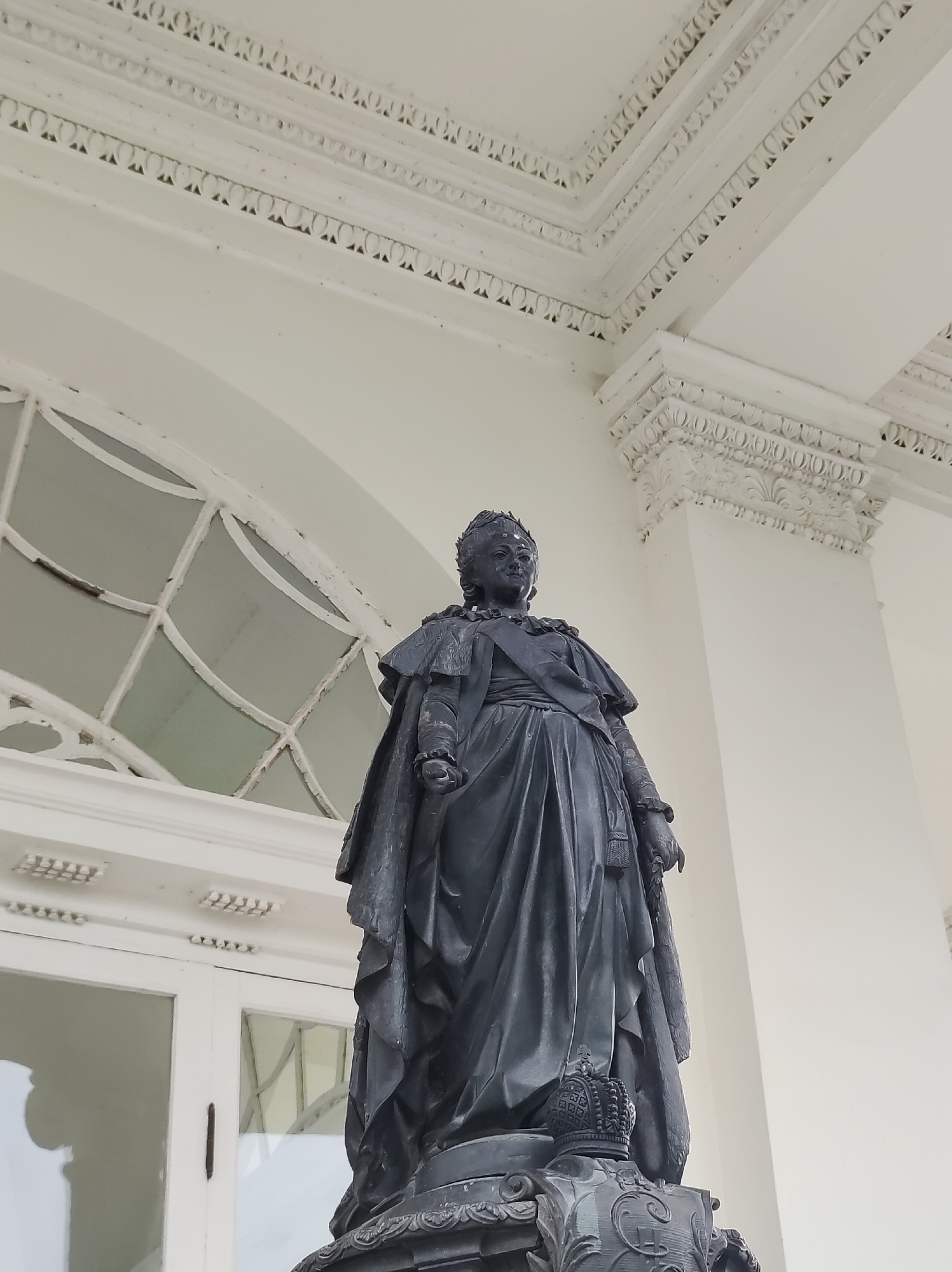 The photograph captures a bronze statue of a woman in regal attire, possibly an empress, standing on a pedestal. The statue is dark in color, with intricate details on the clothing, including a draped cape and a crown on the head of the figure. The pedestal is also ornate and appears to be made of metal.
Behind the statue, a white building with classical architectural elements is visible. The building features decorative molding along the top, a large arched window with a diamond-shaped pattern in the glass, and a white door with glass panes. The overall setting suggests a public space in a city with a historical or grand aesthetic.
The image is specifically located in the city of Pushkin, Russia. The architectural style and the presence of a formal statue are consistent with the historical palaces and grounds found in this city, known for its imperial heritage. The lighting suggests it is daytime, with soft light illuminating the scene.