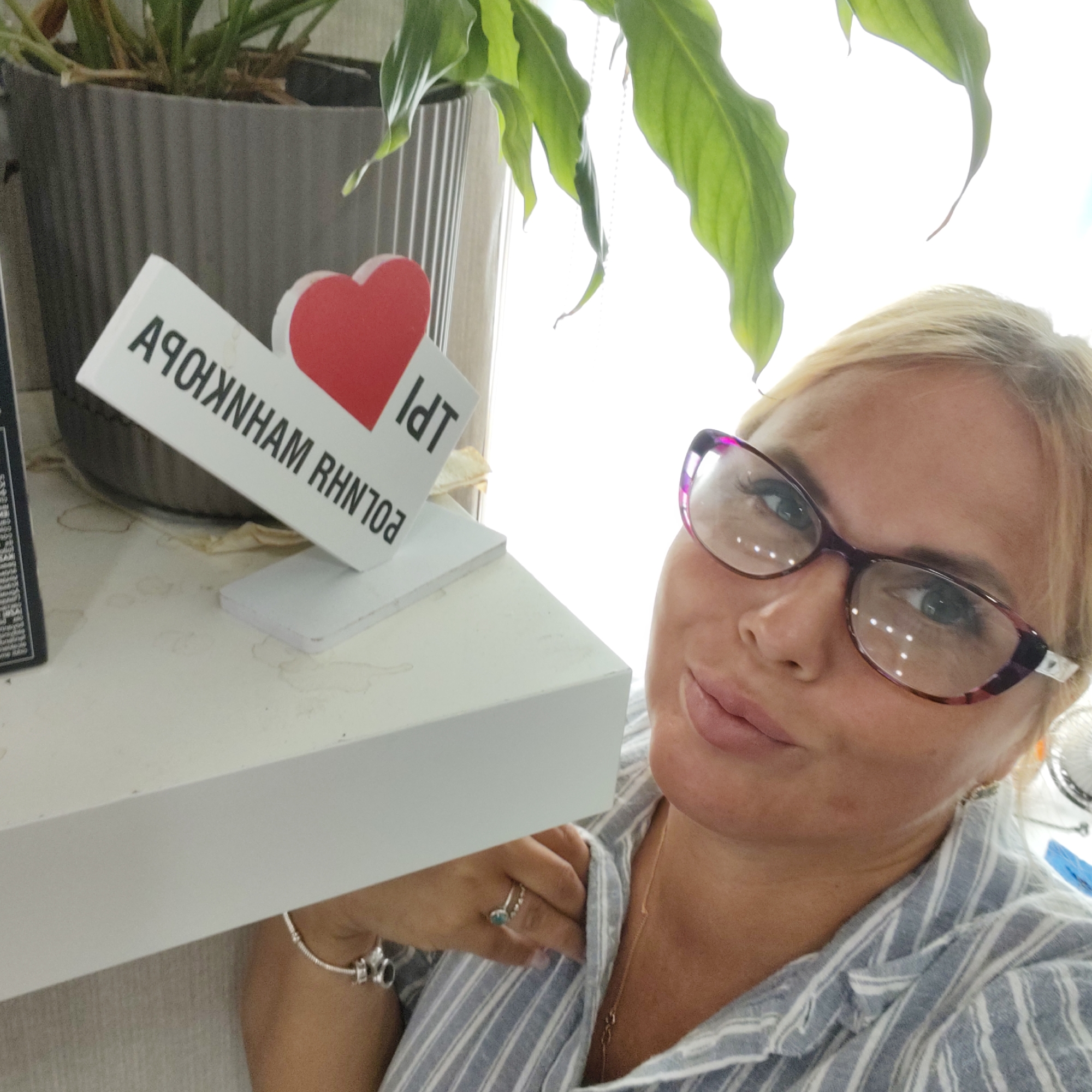 A close-up, selfie-style shot shows a fair-skinned blonde woman with her head tilted to the right, looking at the camera. She has on purple-rimmed glasses and is wearing a light blue and white striped shirt. Her left hand is visible, adorned with a silver bracelet and a ring with a turquoise stone. The background is out of focus, but includes a large green plant in a grey pot, a white shelf, and a brightly lit window. Text on a white object in the foreground reads "I D T" and "АЧОЯХNHAM RHN703". The woman appears to be in an indoor setting.