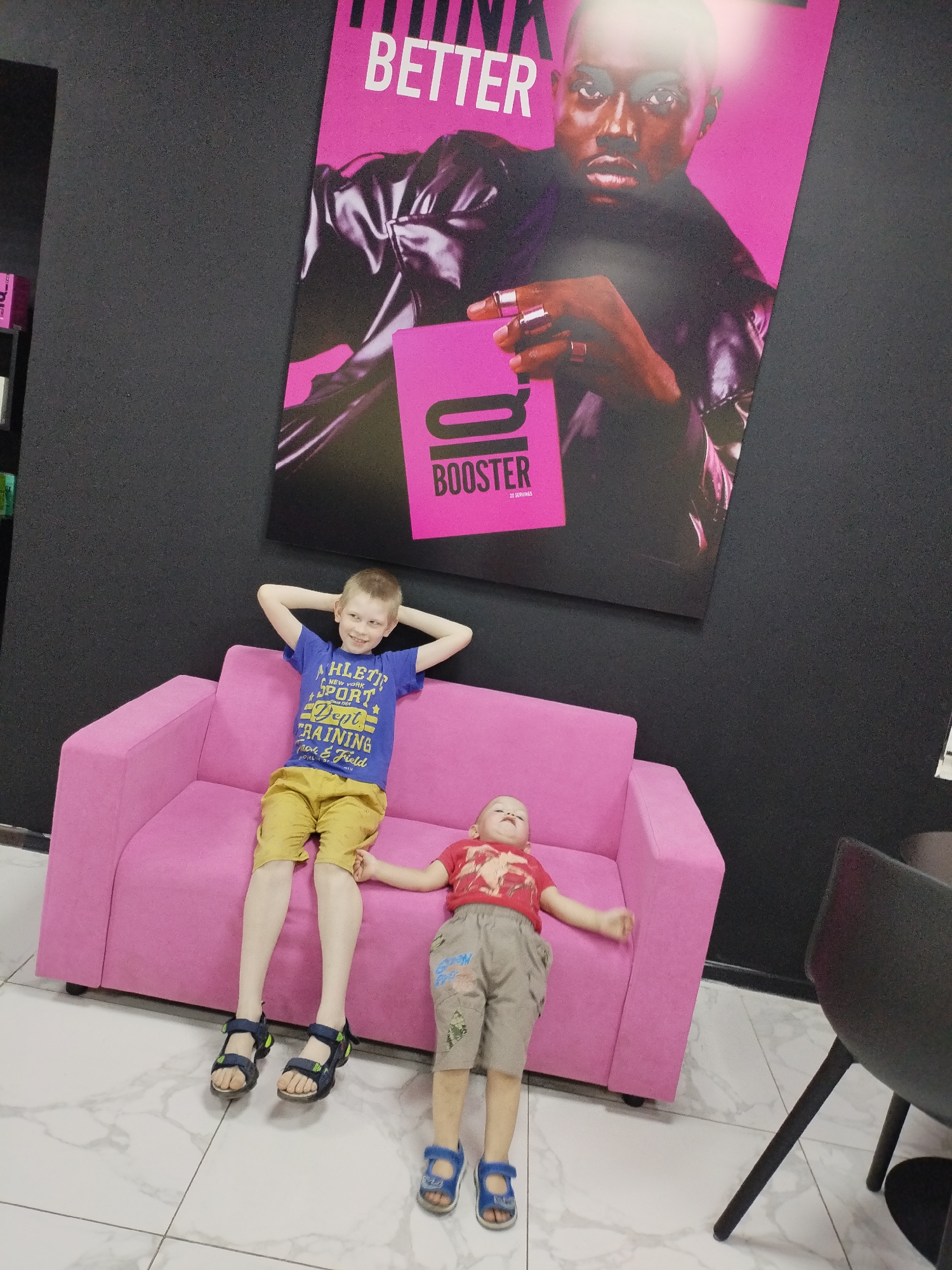 Two young boys are on a pink couch in front of a black wall with a large poster. The boy on the left is sitting up, with his hands behind his head, wearing a blue t-shirt and yellow shorts. The boy on the right is lying down, wearing a red t-shirt and tan shorts. The poster features a man's face and the word "BETTER".