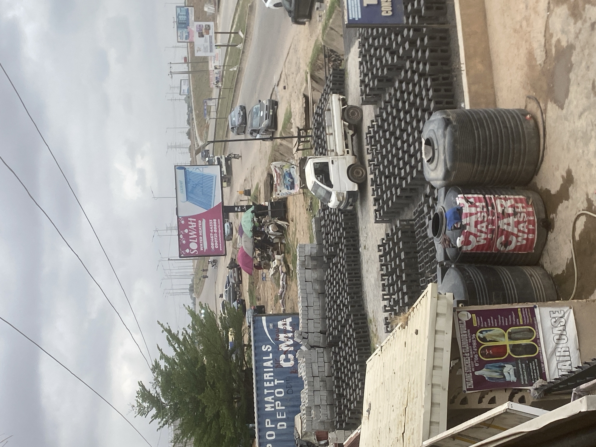 This media file captures a busy roadside commercial area in Lugbe, Nigeria, under an overcast sky during daytime. The scene is dominated by a depot for building materials, with a main road running across the middle ground.
In the foreground and to the right, large stacks of grey concrete blocks are neatly arranged on a dirt ground, indicating a construction supply business. Several black plastic water tanks, some with labels partially visible as "WAS CAR WASH," are also present. A white pickup truck is parked among the stacks of blocks. Adjacent to this area, a large blue shipping container is visible, bearing the text "POP MATERIALS DEPOT CMA" in white lettering. Near the water tanks, a sign in purple advertises "ENLONE STICHES & GLAMOUR WORLD," with a phone number (07067498036) and the slogan "YOUR FASHION & BEAUTY IS OUR PRIORITY," alongside "TAIR CASE."
The main road, with a dusty shoulder, features several cars and a motorcycle. On the left side of the road, a large billboard with a pink background advertises "SOLWAH WATER HEATER" along with phone numbers: "081467-44333" and "090721-88888." Another billboard further down the road displays blue corrugated sheeting. Power lines stretch across the upper left portion of the image against the cloudy, grey sky. A green leafy tree stands on the left, near the road. The overall impression is an active, utilitarian commercial zone.