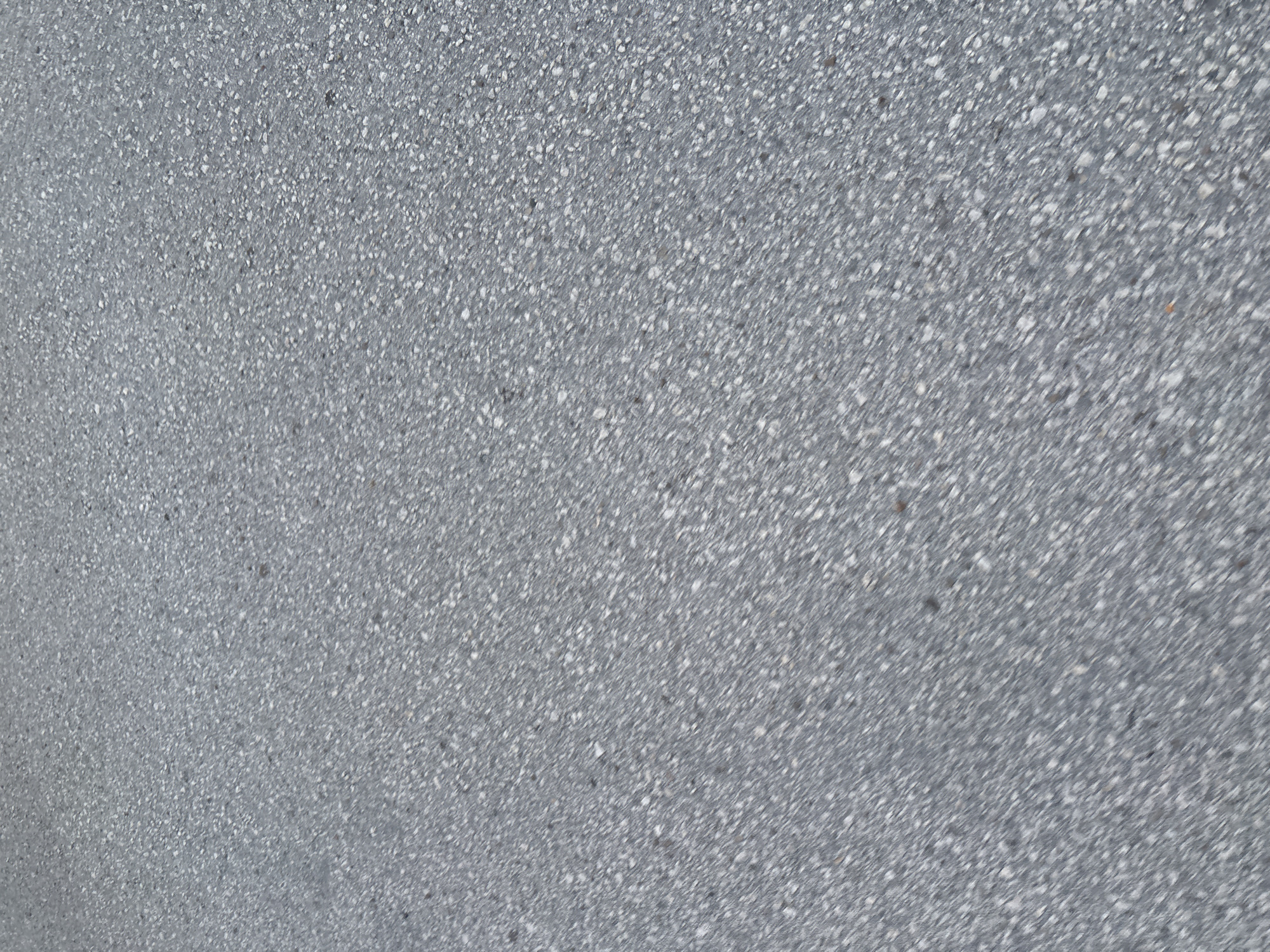 The image displays a close-up, overhead view of asphalt pavement. The surface is textured with small, irregular aggregate stones embedded in a darker matrix. The color palette is predominantly gray, with variations in shade due to the individual stones and lighting. There are no discernible objects, people, or actions present. The scene is devoid of any contextual clues that would indicate its specific location within the city of Plovdiv, Bulgaria.
