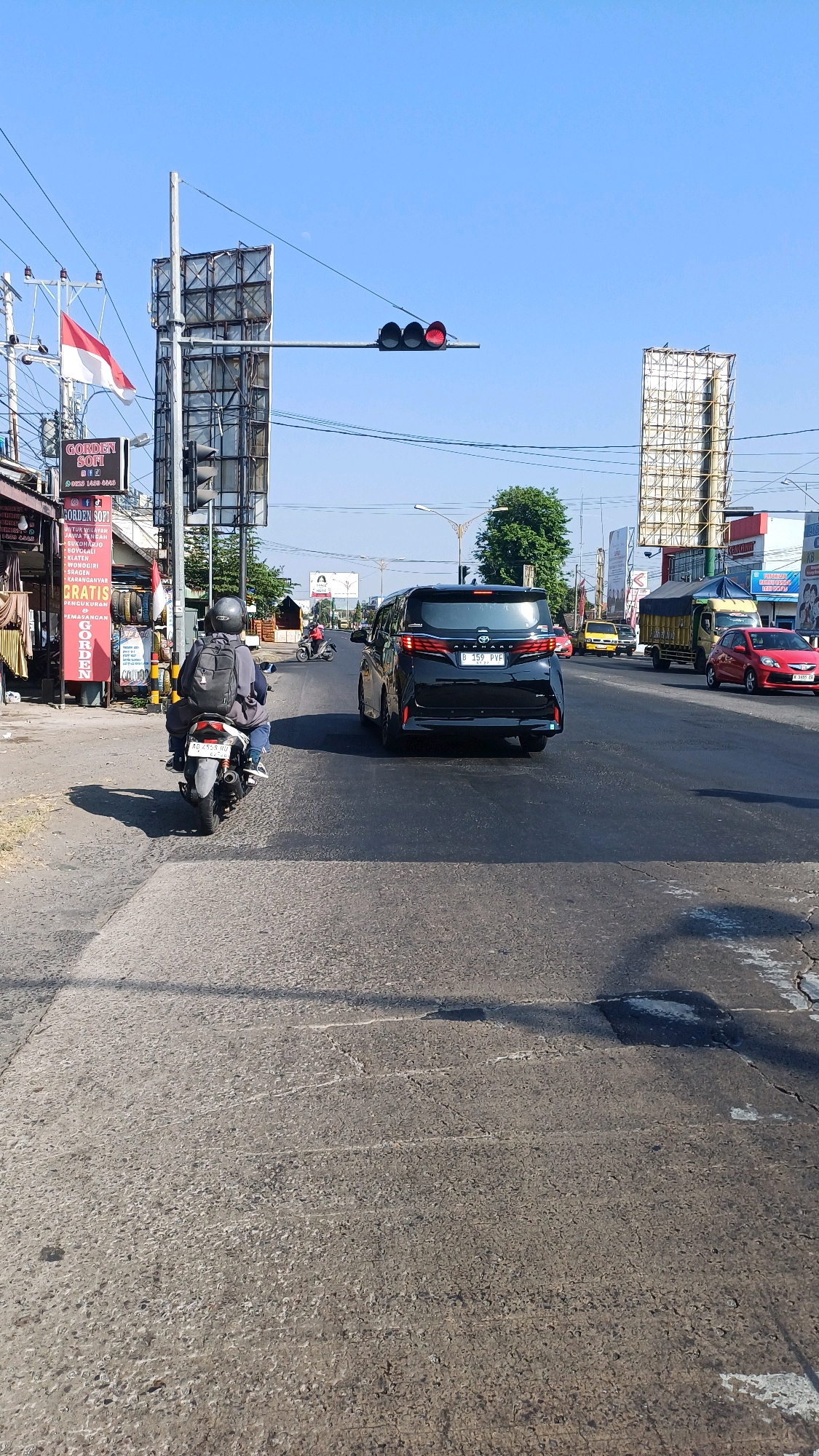 The image captures a street scene in Ngadirejo, Indonesia, on a clear day with a bright blue sky. In the foreground, the asphalt road shows signs of wear, with cracks and patches. A motorcycle, ridden by a person wearing a helmet and a backpack, is in the left lane, moving away from the viewer. To the right of the motorcycle is a black minivan, also traveling in the same direction.
Further down the road, other vehicles are visible, including a yellow car, a truck with a blue tarp, and a red car. Traffic lights are present, with the red light illuminated, indicating a stop. On the left side of the road, buildings line the street, with signs advertising services and products, including a prominent red and white Indonesian flag on a pole. In the background, tall billboards are erected, and trees are scattered along the roadside. The overall impression is a typical urban street during daylight hours.