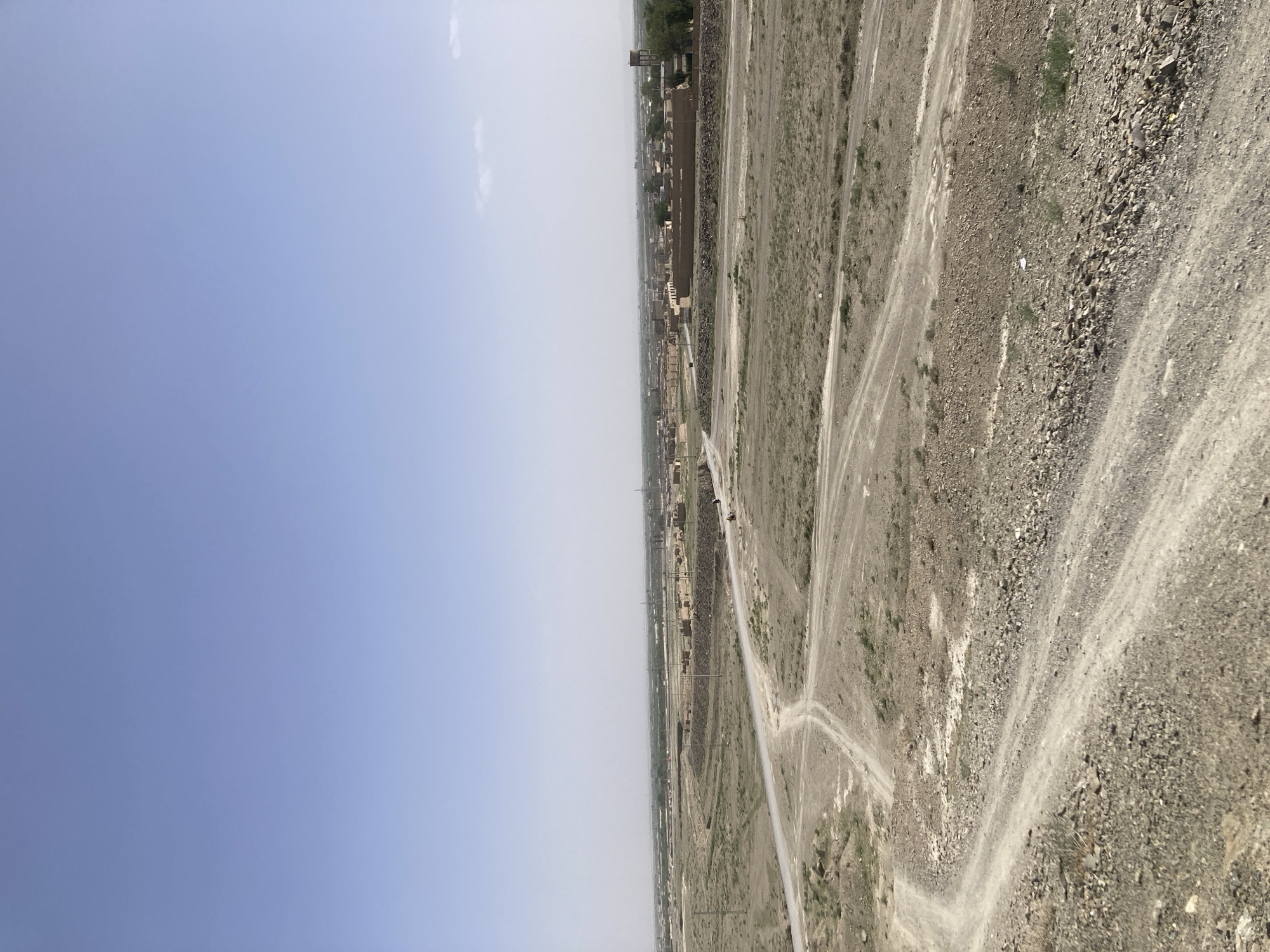 The image depicts an aerial view of Gulistan, Pakistan. The landscape features a flat, arid expanse of land with sparse vegetation and a network of dirt roads. A settlement is visible in the distance, consisting of low-rise buildings and infrastructure. The sky is pale blue with a few wisps of clouds. The overall scene suggests a rural or semi-urban environment. No people are visible.
