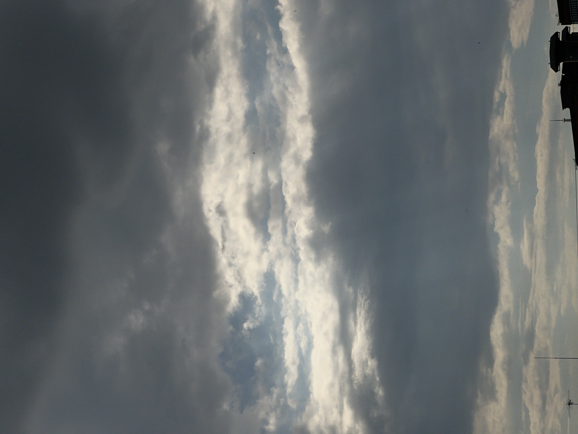 The image depicts a cloudy sky with a sliver of bright light peeking through a break in the clouds. The sky is predominantly gray, indicating overcast conditions. Buildings and power lines are visible in silhouette against the sky. Based on visual references, the location is potentially Krasnodar, Russia.