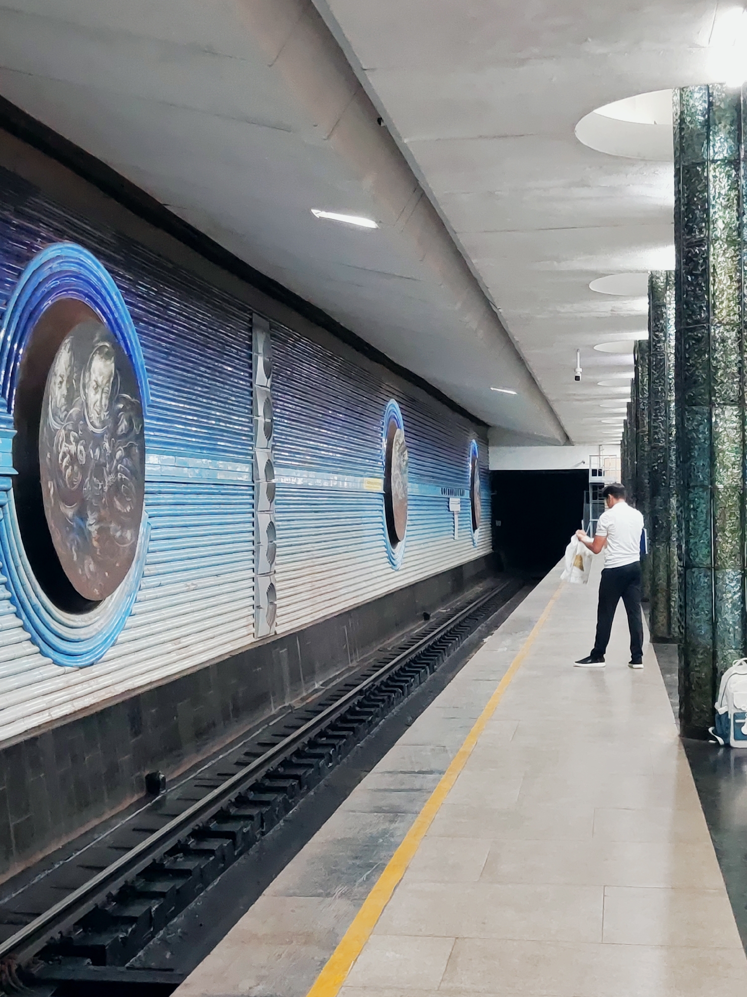 The image depicts a person standing on the platform of a subway station in Tashkent, Uzbekistan. The station features a distinctive mural along the wall, with circular insets that appear to depict astronauts. The platform is lined with a yellow safety strip, and tracks for a train are visible. The ceiling of the station has circular recessed lighting. The person, dressed in a white polo shirt and dark pants, is holding a white plastic bag. To the right, several decorative columns clad in a green, textured material are present, along with a white backpack. The overall impression is of a well-maintained, architecturally interesting public transportation hub.