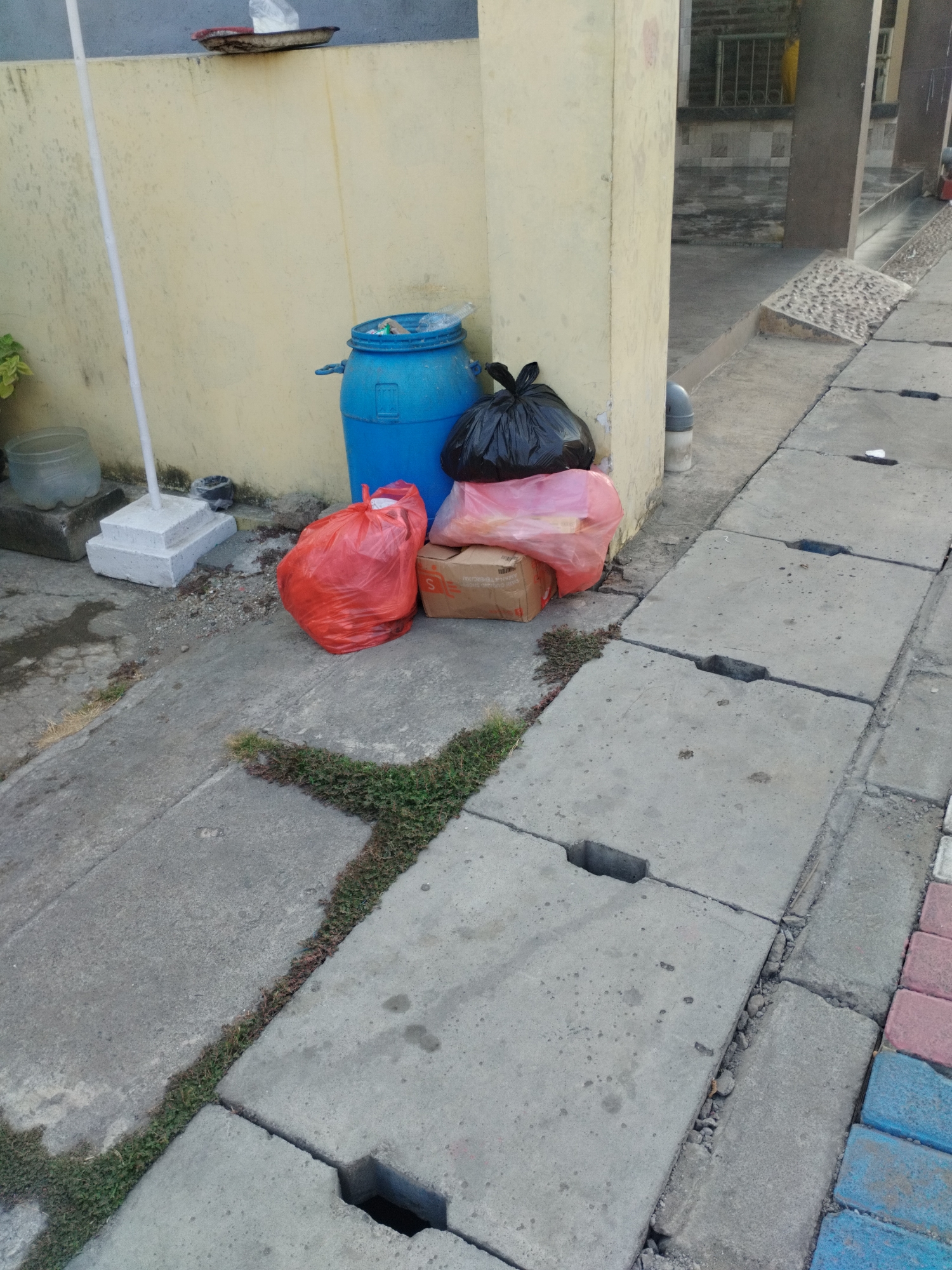 The image depicts a collection of garbage bags and a blue barrel situated on a concrete pathway next to a light yellow wall. The garbage includes two red bags, a pinkish translucent bag, and a black bag. A cardboard box is also present. The pathway shows signs of wear and tear, with patches of grass growing through cracks. The scene appears to be an outdoor area, possibly an alleyway or the side of a building, within the city of Surabaya, Indonesia. The lighting suggests it is daytime.