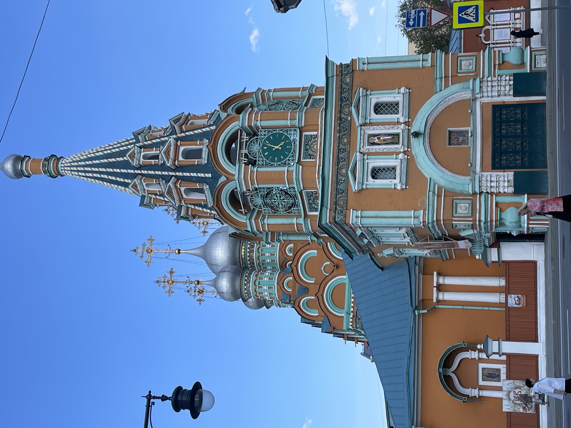 The image shows the exterior of the Church of the Resurrection in Kadashi, Moscow, Russia. The building has a distinct orange and turquoise color scheme, with multiple domes topped with golden crosses. A clock is visible on one side of the building. A street lamp is situated in the foreground. A street sign is placed to the top of the building. Pedestrians are partially visible on the street. The sky is clear and blue.