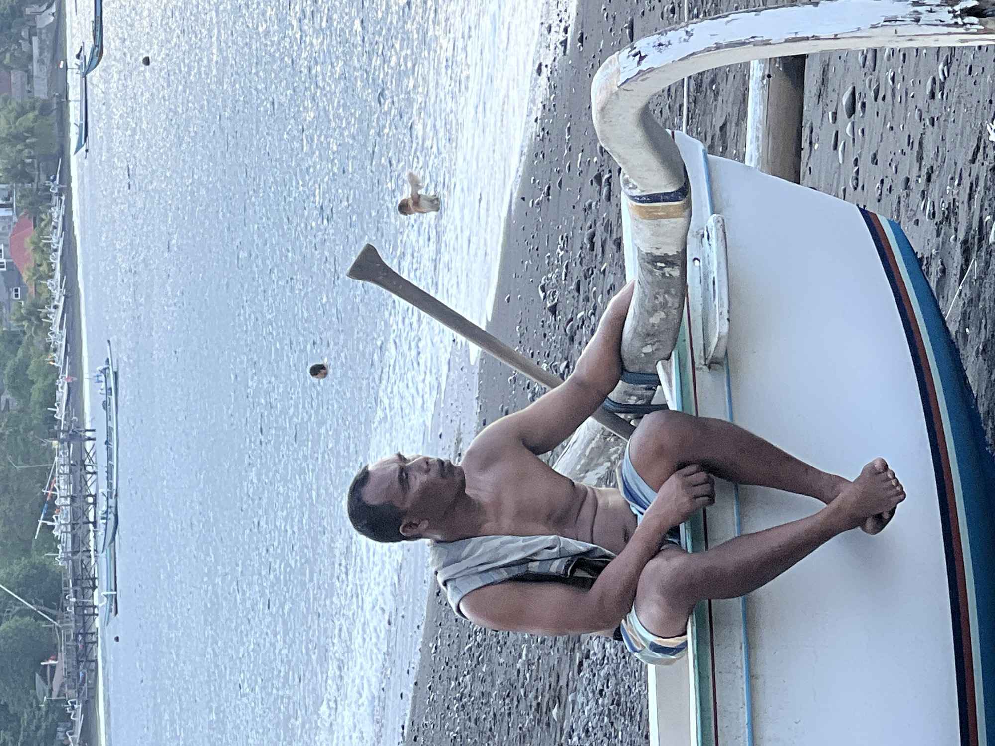 The image captures a man sitting on the edge of a white boat on a rocky beach. The man is bare-chested, wearing striped shorts, and has a cloth draped over his shoulders. He is looking off to the right, with his left arm resting on the boat's outrigger. In the background, the ocean is visible with several boats anchored and a few people swimming. The coastline features a pier with more boats and some buildings nestled among trees. The scene appears to be in Abang, Indonesia, with the overall atmosphere suggesting a coastal fishing village setting.