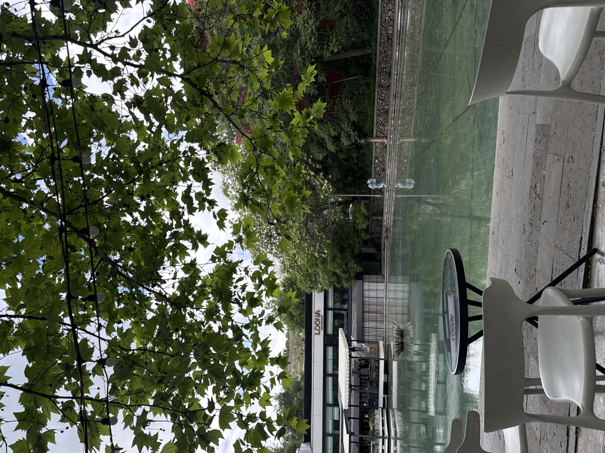 The image shows an outdoor cafe scene in Yerevan, Armenia. A tree with bright green leaves and strings of lights hangs overhead. In the background is a building labeled "LOONA" with a modern architectural design. A body of water, possibly a pond or a fountain, reflects the building and the trees. Several white plastic chairs and a round table are situated on a paved area, partially visible on the right side of the frame. No people are readily visible in the foreground.