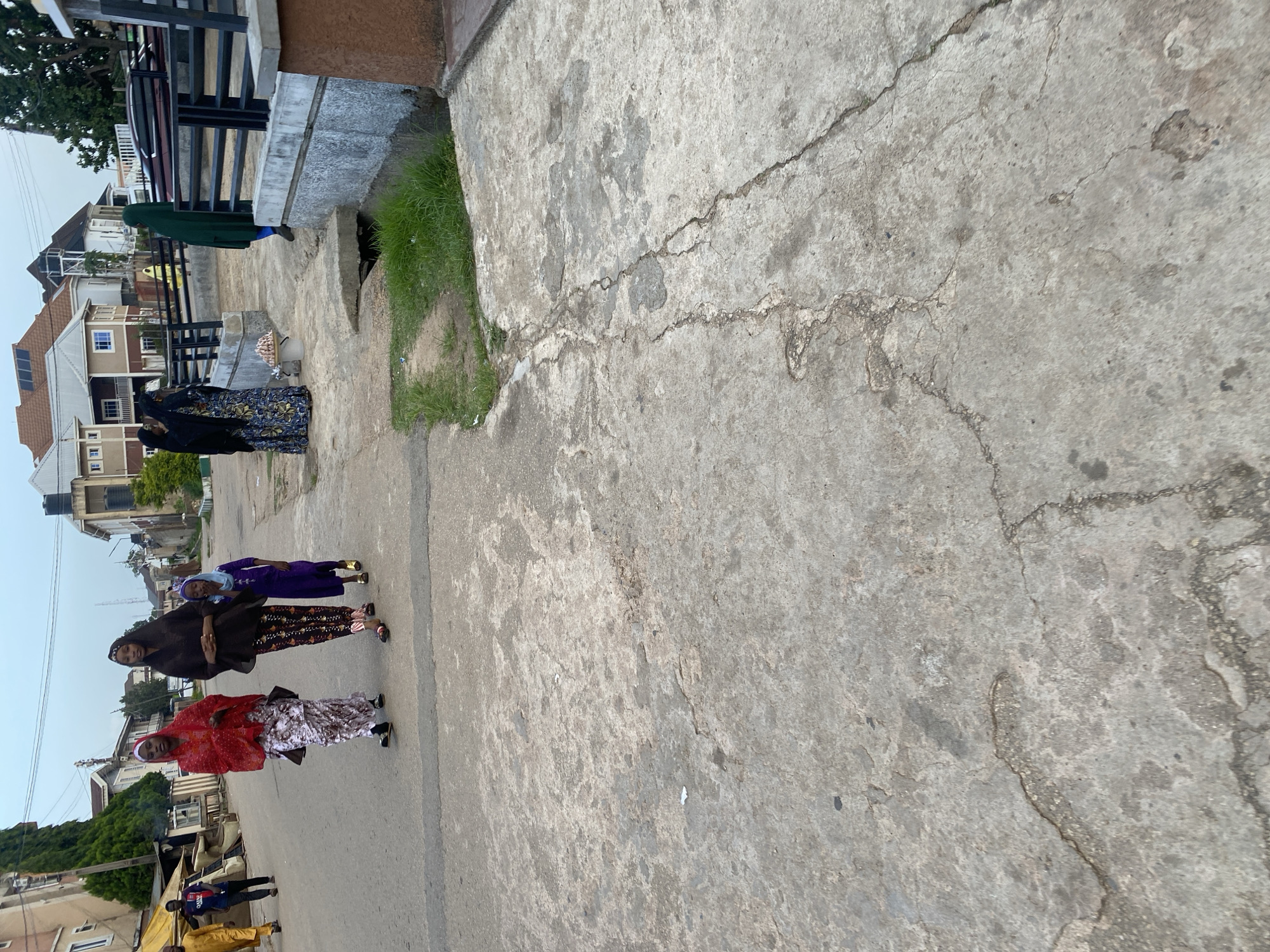 In Jos, Nigeria, a group of people are seen walking on a paved street. In the foreground, the cracked concrete walkway takes up most of the frame. Further down the street, three women and a young girl are walking together. The woman on the left wears a red hijab and a patterned skirt, while the woman next to her has a brown hijab and patterned trousers. The young girl in the middle is dressed in a purple abaya. To the right, another woman is visible wearing a green hijab. In the background, residential buildings and lush green trees line the street, suggesting a suburban or residential area.
