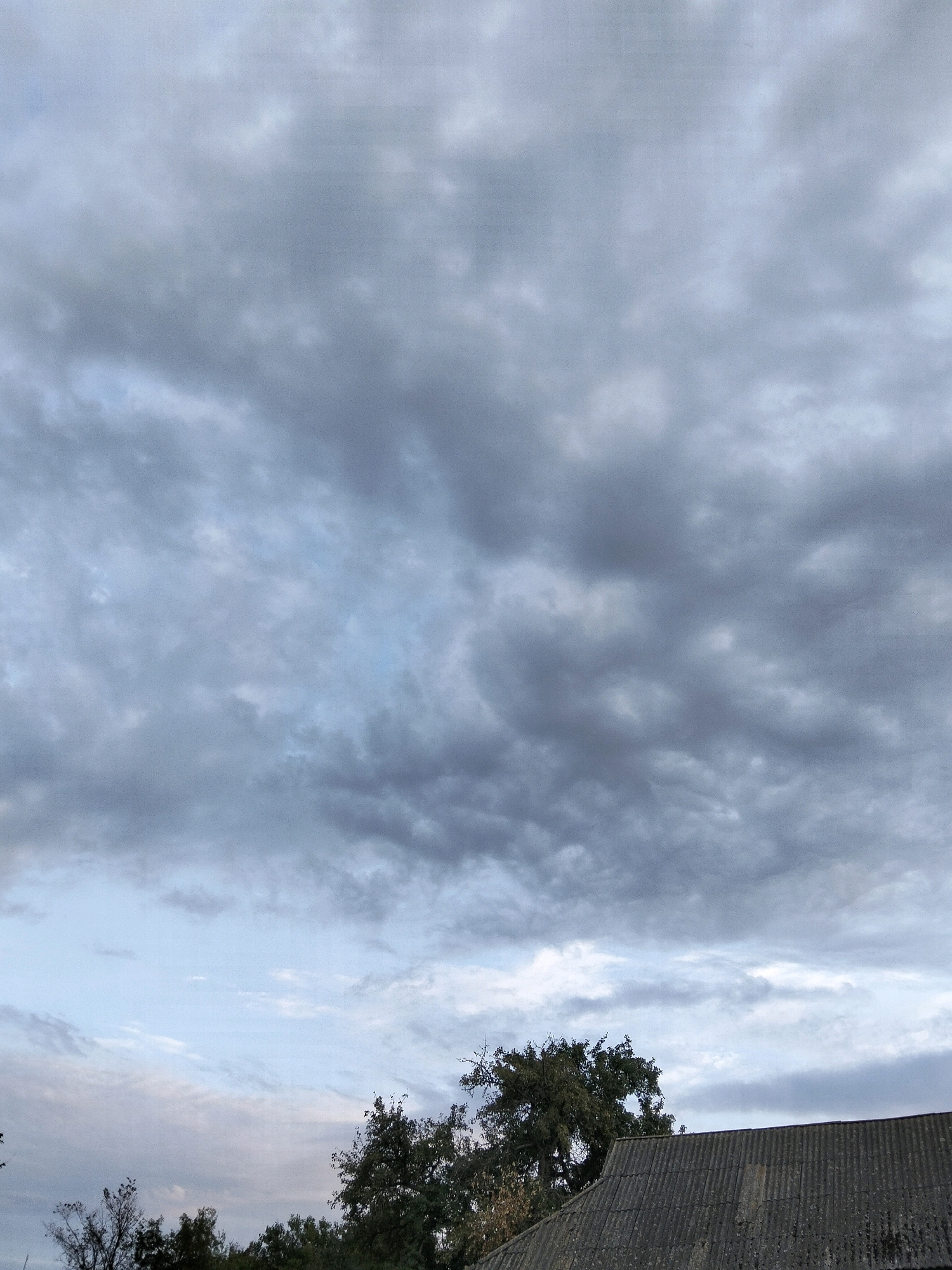 The image shows a cloudy sky above a tiled roof and trees in Ustyvytsia, Ukraine. The sky is filled with dark gray clouds, with some lighter patches of blue sky visible. The roof is made of corrugated material, likely slate or asbestos. The trees are full of dark green leaves. The overall impression is one of a gloomy, overcast day.