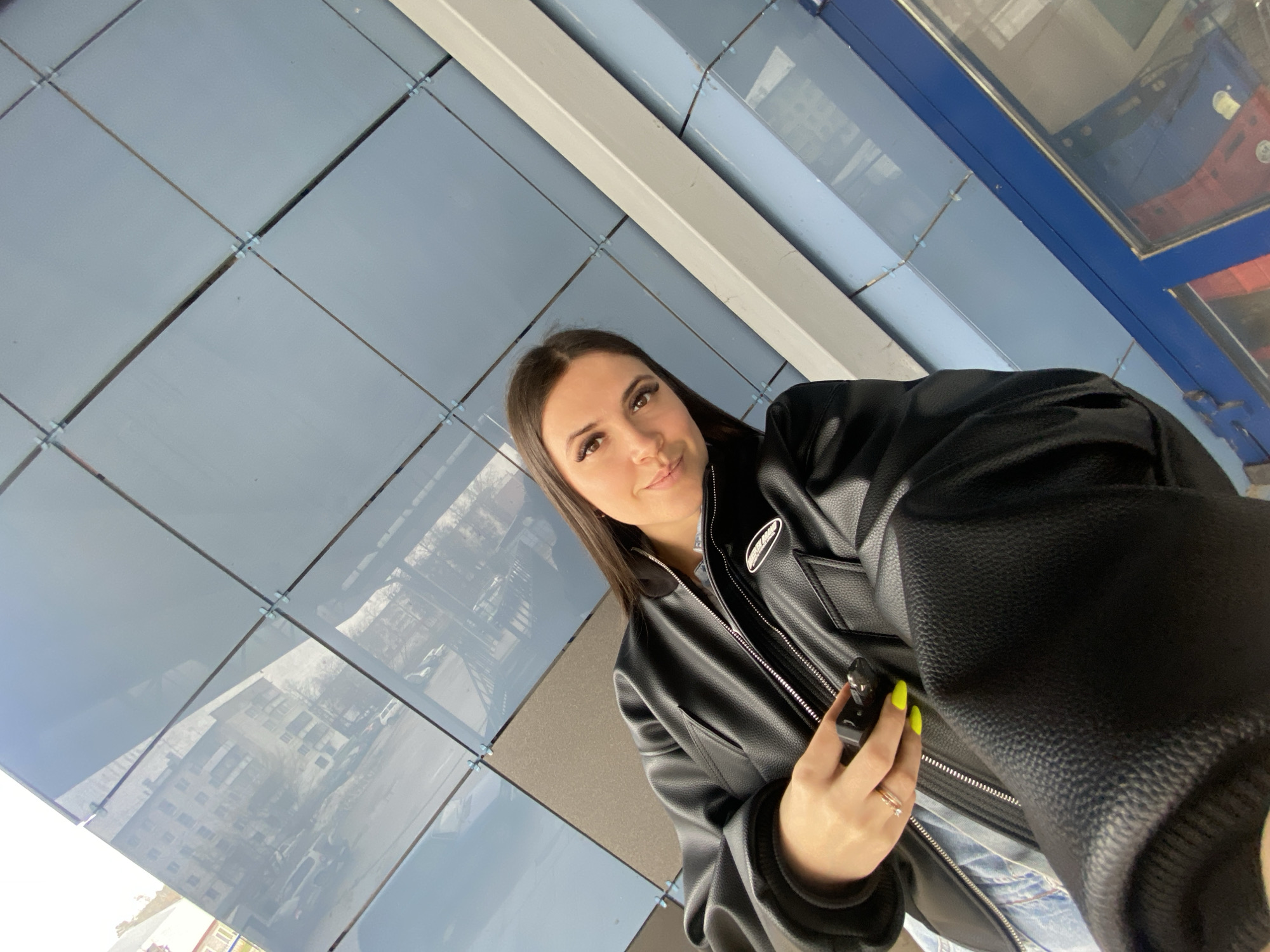 A woman is taking a selfie in Chaikovsky, Russia. She has long brown hair, fair skin, and wears a black leather jacket. The location appears to be an enclosed walkway or shelter with a glass ceiling composed of individual square panes. Reflections of city buildings are visible on the glass. The woman is holding a small, dark object, possibly keys or a remote. Her fingernails are painted bright yellow.