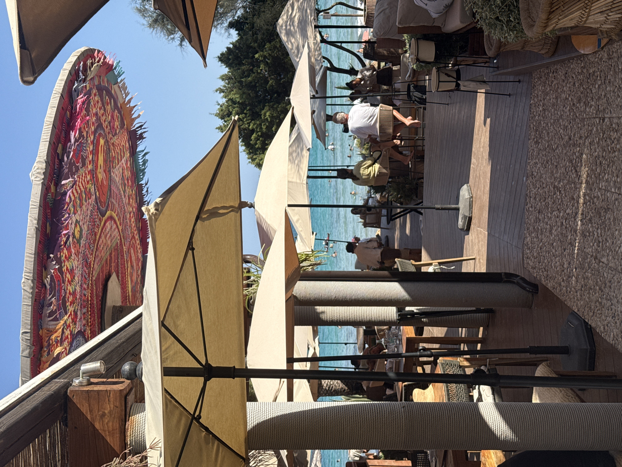 The image depicts a beachfront restaurant in Santa Eulària des Riu, Spain. Several large, cream-colored umbrellas provide shade over an outdoor seating area. In the background, the turquoise waters of the Mediterranean Sea are visible, with a few people swimming and boating. The restaurant features wooden decking and furniture, with various patrons seated at tables and on stools. The decor includes woven baskets and potted plants. A vibrantly patterned, fringed parasol hangs overhead, adding a bohemian touch to the scene. The atmosphere appears relaxed and sunny, typical of a Mediterranean coastal setting.
