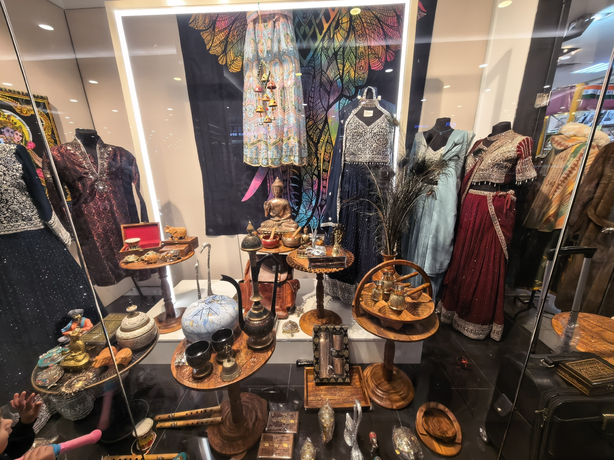 The image depicts a storefront window display in Moscow, Russia. The display features mannequins dressed in colorful Indian clothing. Various decorative items and artifacts are displayed on wooden stands and trays. The backdrop includes a large elephant-themed graphic. Reflected in the glass are elements of the surrounding street. A child's hands are visible in the lower-left corner, indicating someone is standing nearby viewing the display. A suitcase is visible in the reflection in the right side of the image.