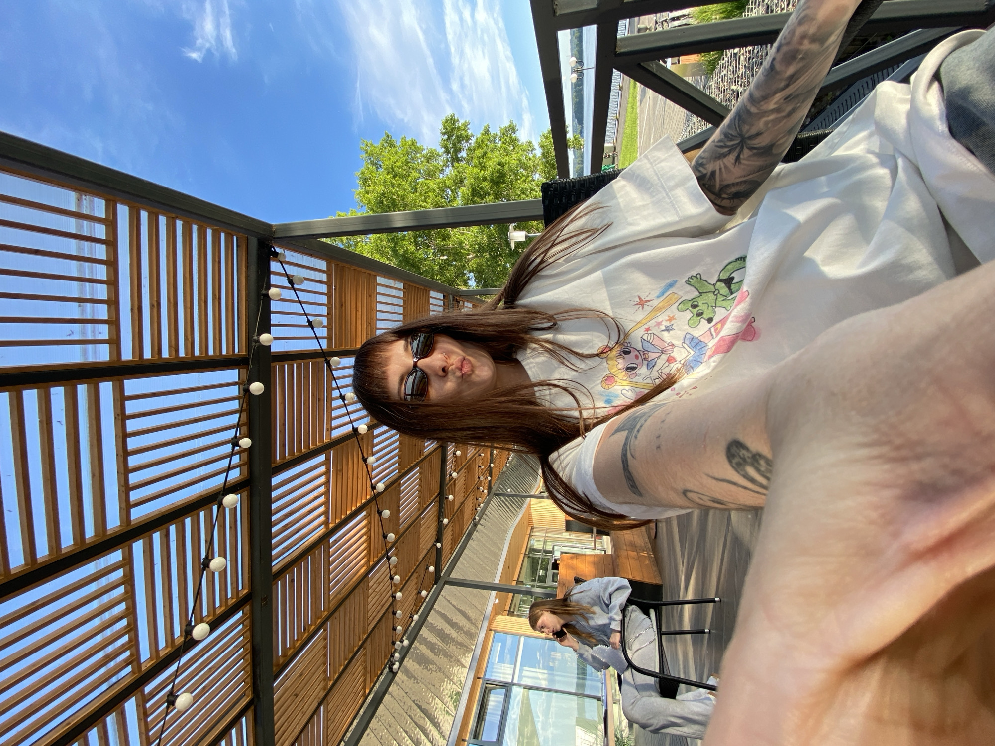 A brightly lit selfie captures a young woman seated outdoors under a modern pergola structure. The image is taken from a low angle, showing the woman's face in the foreground, turned slightly to her left, with her lips pursed in a playful kissy face. She has long, dark hair with bangs and wears dark sunglasses. Her white t-shirt features colorful cartoon graphics, including what appears to be characters like Sailor Moon and a green creature. Both of her visible arms are heavily tattooed, with one arm displaying a design that includes palm trees.
Behind the foreground subject, another young woman in a grey sweatsuit is visible, sitting at a black table and looking down at her phone. The setting is an outdoor patio or cafe, characterized by an overhead wooden slat roof supported by dark metal beams. String lights with spherical bulbs are strung across this ceiling structure, adding to the ambiance. In the background, beyond the seating area, a modern building with large reflective windows can be seen, along with vibrant green trees under a clear blue sky dotted with wispy white clouds. The bright sunlight indicates it is daytime, likely a sunny day. No discernible text is visible on clothing or in the surroundings.
