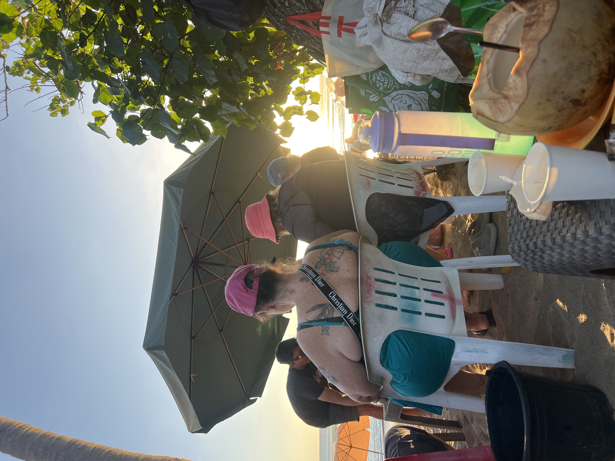 At a Kuta, Indonesia beach during sunset, two women are seated on plastic chairs under a large green umbrella. The woman in the foreground, facing away from the camera, wears a pink baseball cap, a teal swimsuit, and has numerous tattoos on her back and shoulders. The woman to her right, also facing away, wears a pink bucket hat and a dark jacket. Behind them, a man in a black t-shirt bends over, possibly attending to something on the sand. The beach environment is indicated by the presence of sand, beach chairs, an umbrella, and the ocean visible in the background. Several items are placed on the ground and on a woven surface, including a black bucket, white cups, a water bottle, shopping bags, and a halved coconut with a straw and spoon. The sun is low on the horizon, casting a warm glow and creating a serene atmosphere.