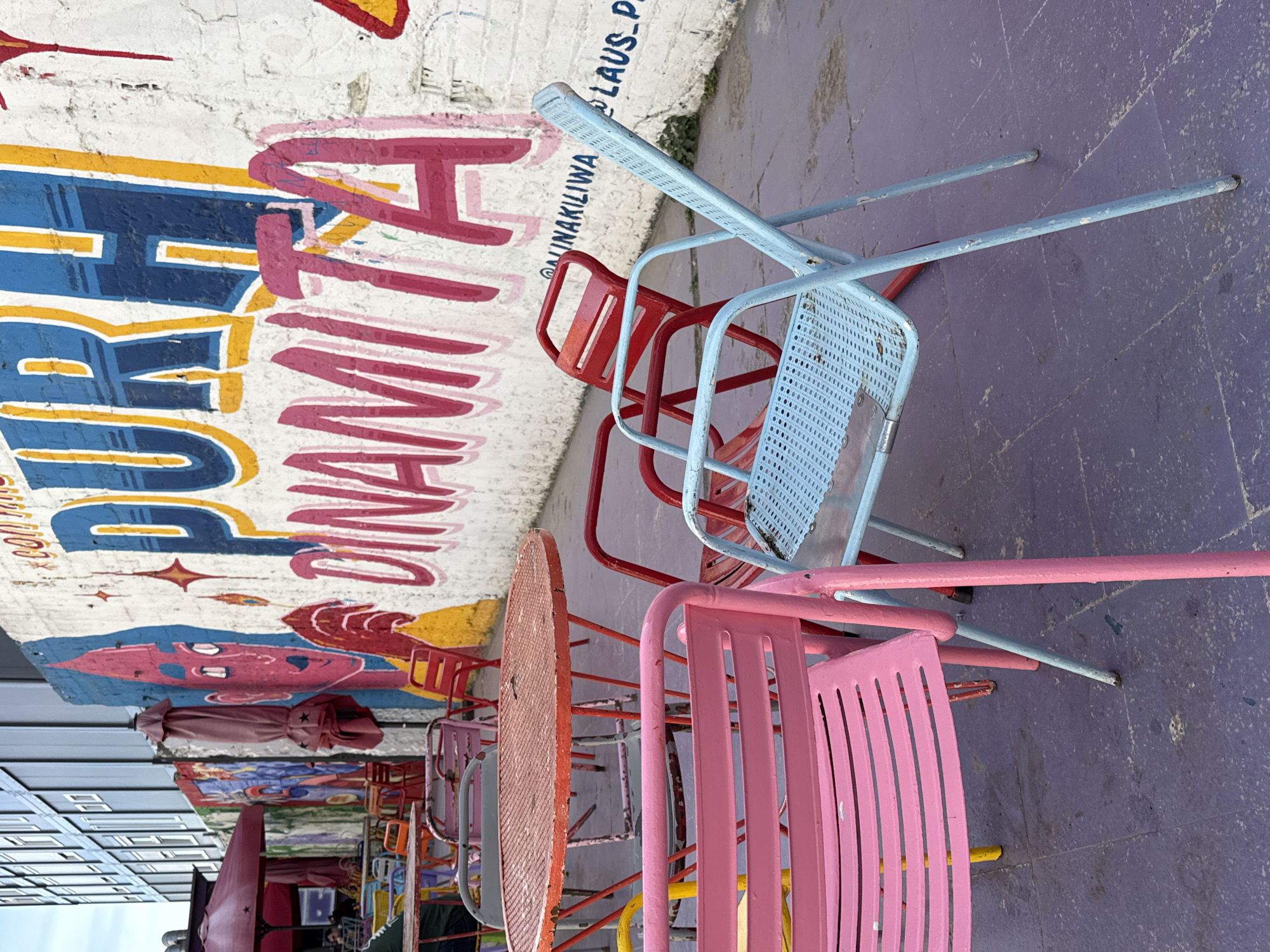 The image depicts an outdoor scene with a mural and colorful patio furniture. The mural, painted on a white brick wall, features a cartoonish face and bold text that reads "DINAMITERS." A variety of metal chairs and tables in different colors (pink, red, blue, orange, yellow) are arranged on a purple-painted pavement. The chairs appear to be stacked or placed haphazardly. The wall is also tagged with "@MINAKILIWA" and "LAUS. P". The image is taken in Barcelona, Spain.