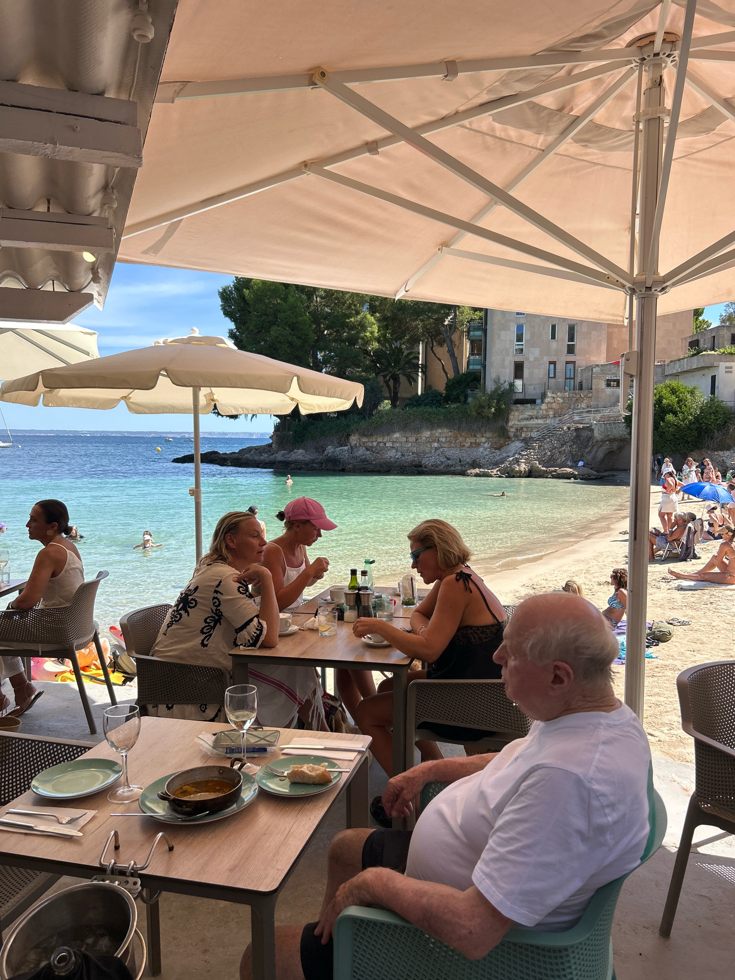 In Cas Català-ses Illetes, Spain, this image captures a scene at an outdoor beachfront cafe under a large cream-colored umbrella. The turquoise Mediterranean Sea forms the backdrop, with a sandy beach populated by sunbathers visible. Four individuals are seated at a table in the foreground, engaged in dining. Two women sit opposite each other at the table, one wearing a white patterned top and the other a black lace top. A man in a white t-shirt is seated facing away from the camera, looking towards the sea. Another person, whose face is obscured, is seated to his right. On the table, there are plates, cutlery, wine glasses, a bowl of food, and drinks. In the background, more people are visible on the beach, some lounging and others walking. Buildings and trees line the coastline behind the beach. The overall atmosphere suggests a relaxed, sunny day at a seaside establishment.