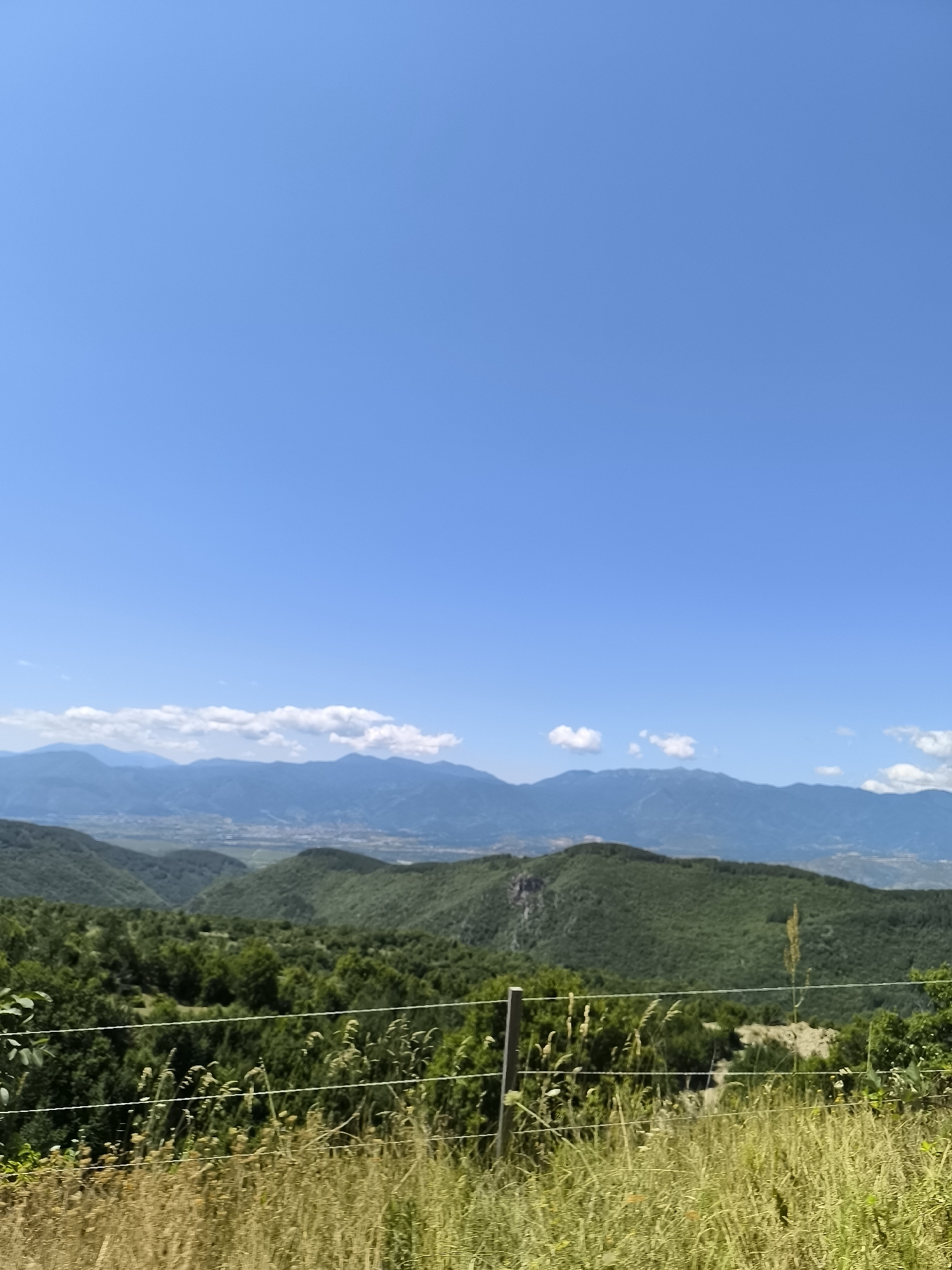 The image captures a panoramic view of a mountainous landscape in the vicinity of Leshten, Bulgaria. The scene is dominated by rolling green hills and mountains under a clear, bright blue sky. In the foreground, a wire fence runs horizontally across the frame, with dry, tall grass and foliage in front of it. The middle ground is covered in lush green trees and dense vegetation, with some areas appearing as forested slopes. In the background, a range of distant, hazy mountains stretches across the horizon, with a hint of a valley or plains with structures visible in the far distance. A few scattered white clouds are present in the sky, adding a subtle texture. There are no people or animals visible in the image. The overall impression is one of a serene and natural environment, likely captured during daylight hours.