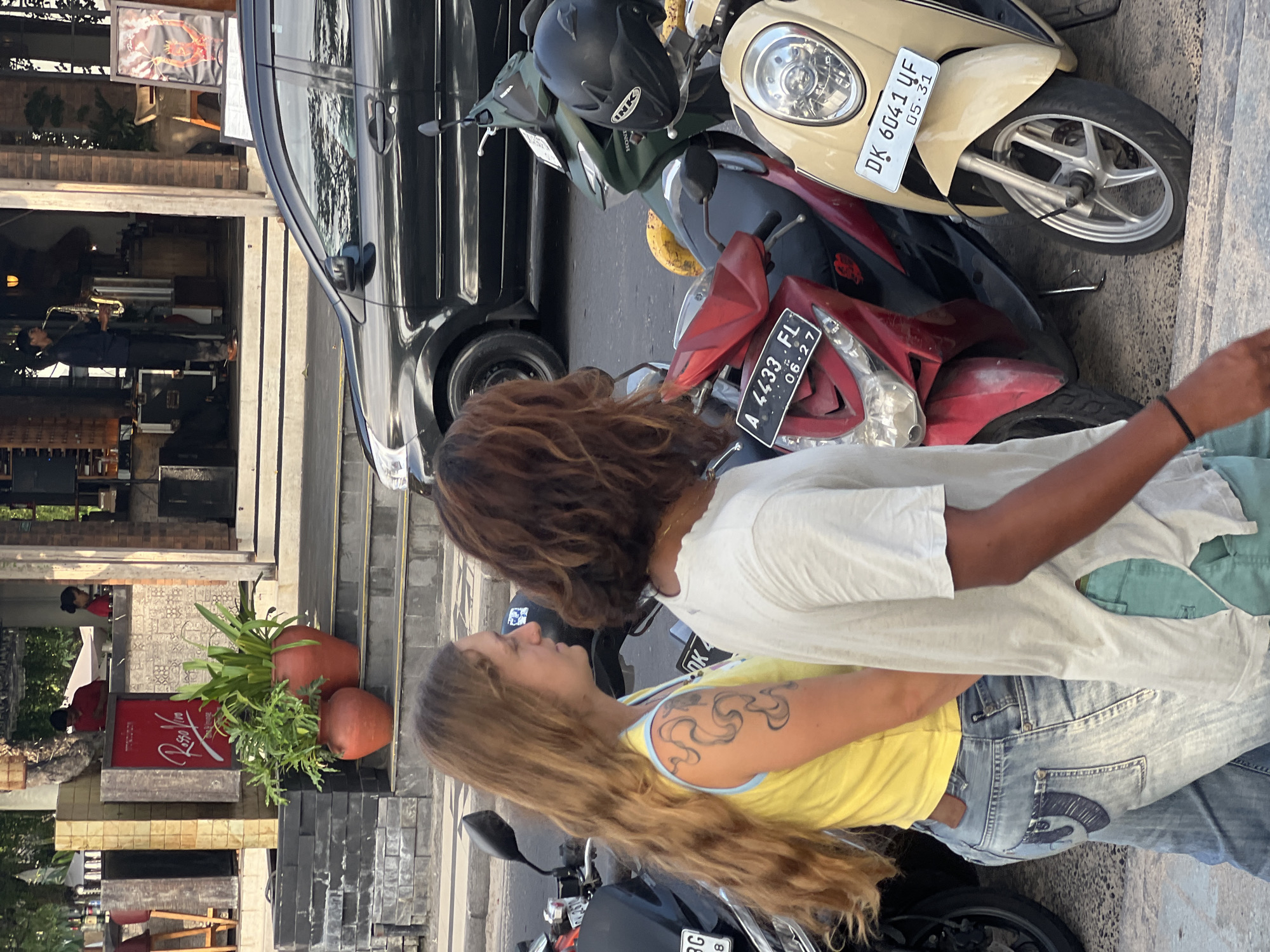 In Kuta, Indonesia, two individuals, a woman with long blonde hair and a man with curly brown hair, walk side-by-side on a paved surface. The woman wears a yellow sleeveless top and light blue jeans, with a tattoo visible on her left arm. The man is dressed in a white t-shirt and teal shorts. Behind them, several parked motorbikes are visible, including a red one with license plate "A 4433.FL" and a beige one with license plate "DK 6041 UF." A black car is parked further behind the motorbikes. In the background, a building with a red sign reading "Rosso Vivo" and planters with greenery are present. A person is seen playing a saxophone on an elevated platform within the building's entrance area.