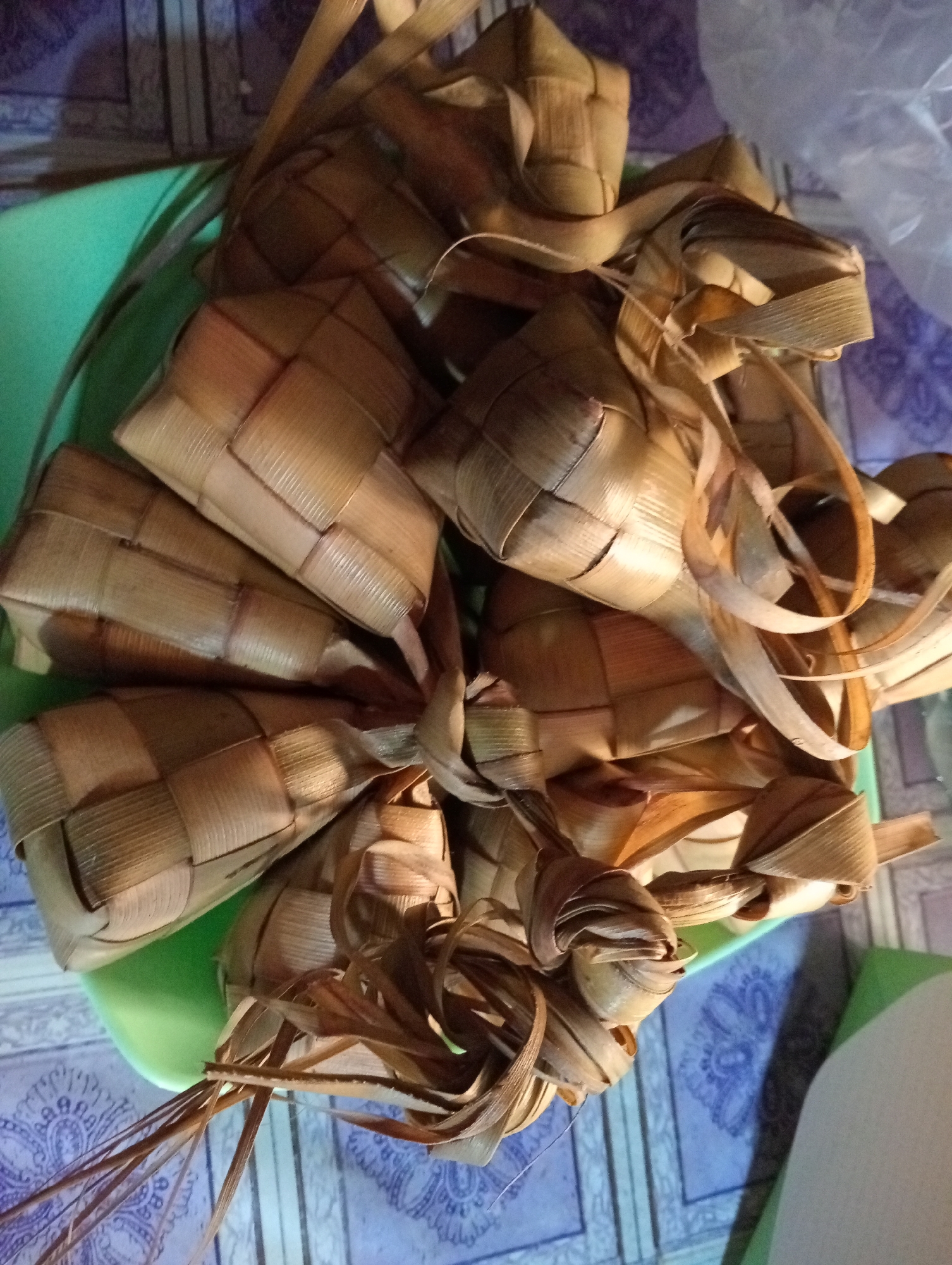 A pile of ketupat, a traditional rice cake wrapped in palm leaves, is displayed on a green surface. The ketupat are arranged in a cluster, with some showing intricate woven patterns. The background consists of a patterned surface, possibly a table or floor, with a plastic bag visible on the right side. The lighting is dim, casting shadows on the objects. The scene appears to be indoors, with the setting likely being Krajan Kidul, Indonesia, as suggested by the context. There are no people or animals visible in the image. The ketupat are the sole focus of the photograph.