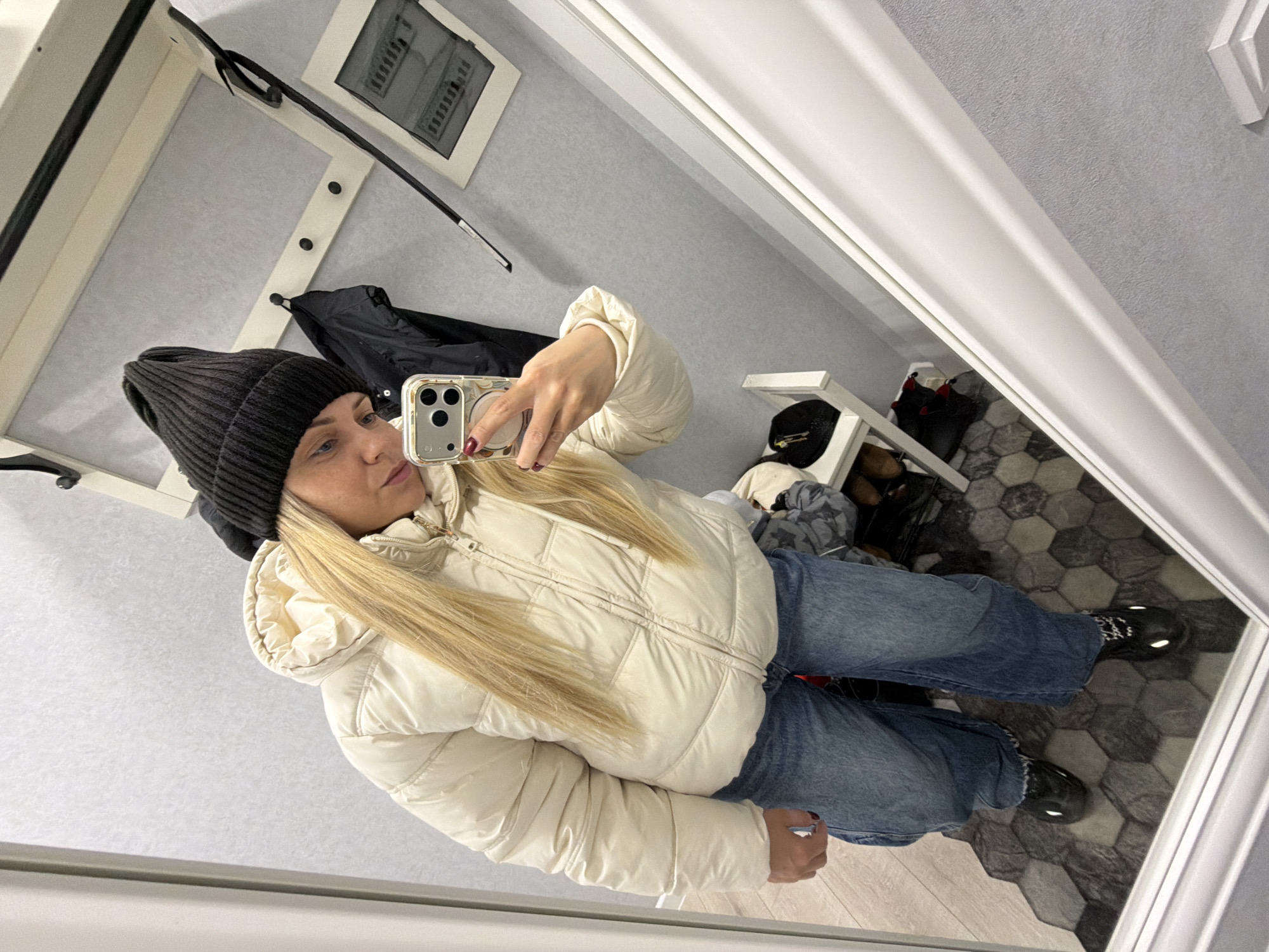 A woman is taking a selfie in what appears to be a dressing room. She is wearing a black beanie, a light-colored puffer jacket, and blue jeans. She is holding a phone with a reflective case. The room has light gray walls and a hexagonal patterned floor. There's a clothing rack and some clothing items visible in the background. There is no information available to discern the exact location, or the city of Votkinsk. The image mainly shows the woman and the immediate surroundings of the dressing room.