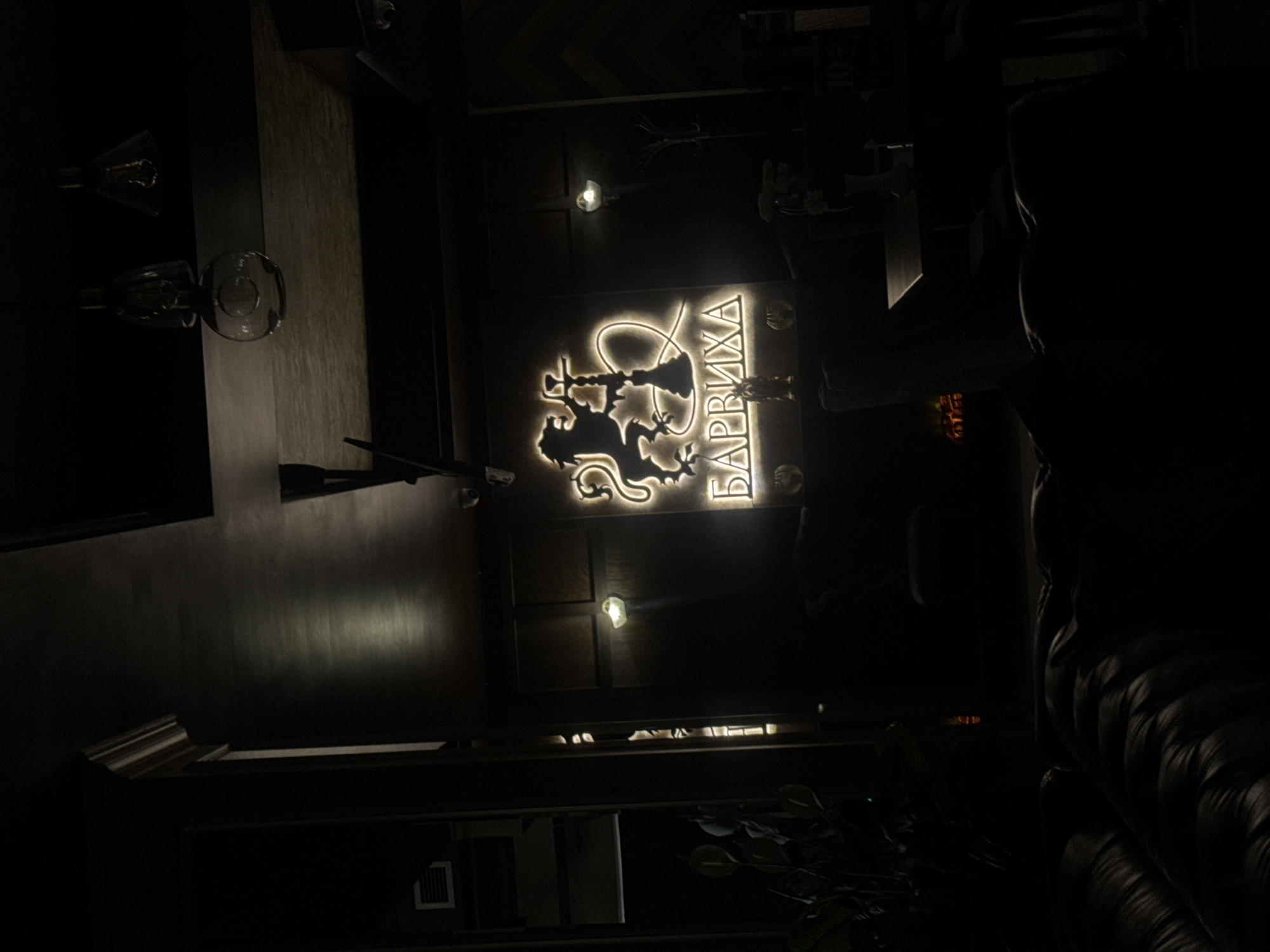 The image depicts the interior of a dark, dimly lit establishment, possibly a hookah lounge or bar. A prominent illuminated sign with a lion and hookah motif is visible, with Cyrillic lettering below it that reads "БАРВИХА" (Barvikha). The sign is the primary light source in the scene. In the foreground, plush dark leather seating is visible, suggesting a seating area. In the background, there are wooden paneled walls and ceiling, with several hanging light fixtures that are not actively illuminating. Some potted plants are present on the left side of the image. The overall ambiance is dark and moody. The specific location is identified as Ussurijsk, Russia. There are no people clearly visible in the image.
