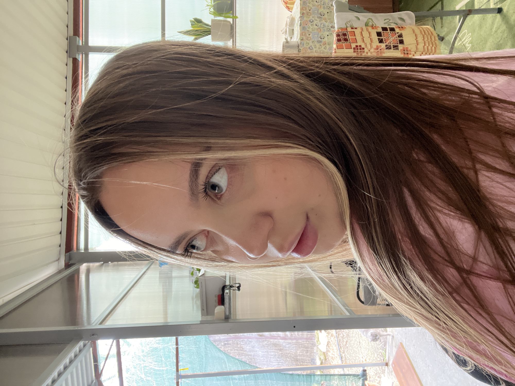 A young woman with brown hair and blonde highlights is looking down and to the left. She is wearing a pink top. The background appears to be a greenhouse or sunroom with translucent panels. Outside the structure, greenery and what may be a yard or garden are visible. There are objects on a shelf inside the structure behind her. The location provided (Novopokrovskaya, Russia) is not possible to confirm, as this cannot be discerned from the image.