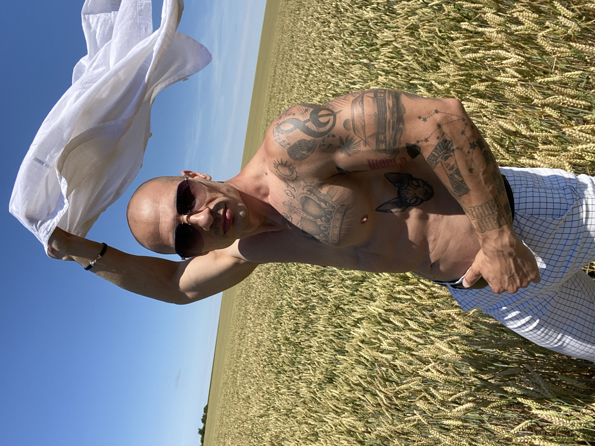 A shirtless, tattooed man wearing sunglasses and checkered pants stands in a wheat field under a clear blue sky. He is holding a white piece of fabric above his head with his right arm. The image was taken in Zgalevo, Bulgaria.
