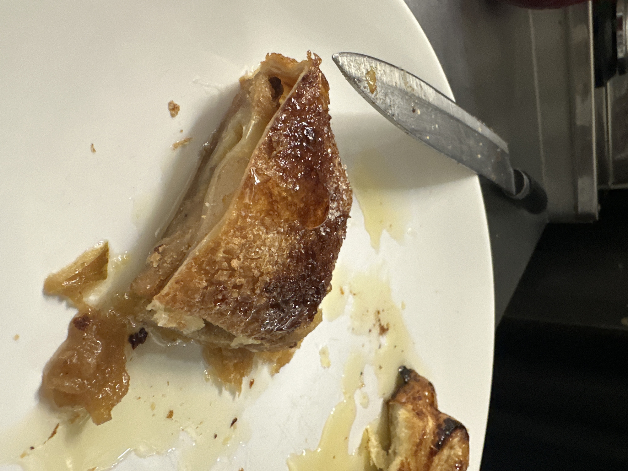 A close-up shot shows a partially eaten pastry dish on a white plate, accompanied by a knife. The pastry is golden brown with a flaky texture. Drippings of liquid surround the pastry remains. The knife has a metal blade and a black handle. The setting is a kitchen environment with stainless steel components visible in the background. There is no evidence of the image being taken in the city of Новоглаголево, Russia. There are no people or interactions visible in the image.
