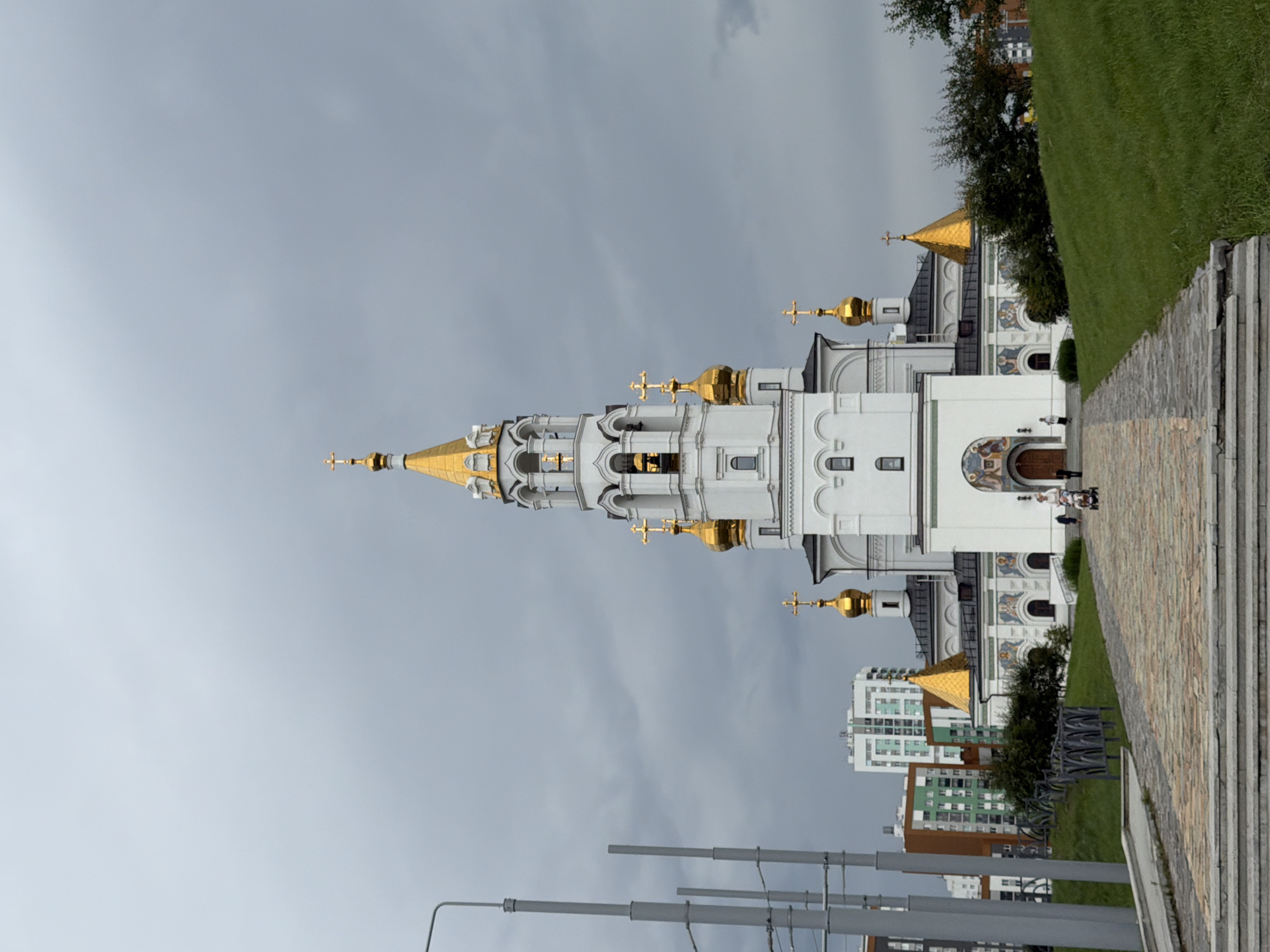 The image shows a white church with golden domes and crosses against a cloudy sky. The church has intricate architectural details, including arched windows and decorative elements. In the foreground, there is a stone pathway leading up to the church, bordered by a grassy slope on the right. Several modern buildings are visible in the background, suggesting an urban or semi-urban setting. A few people are walking on the pathway and near the entrance of the church. The lighting suggests it is daytime, but the overcast sky creates a subdued atmosphere. The image is oriented vertically, with the church tower extending upwards from the bottom of the frame.